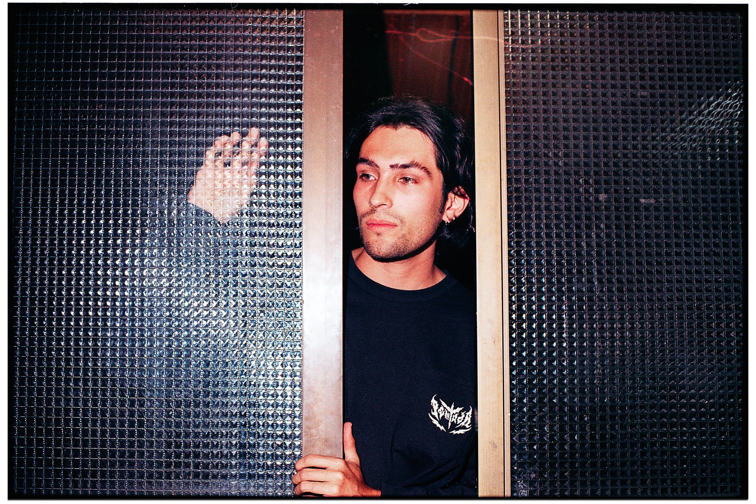 Joven de cabello largo y oscuro asomándose por una puerta de ascensor de metal perforado, con una mano en la puerta y ropa negra con un diseño en la camiseta.