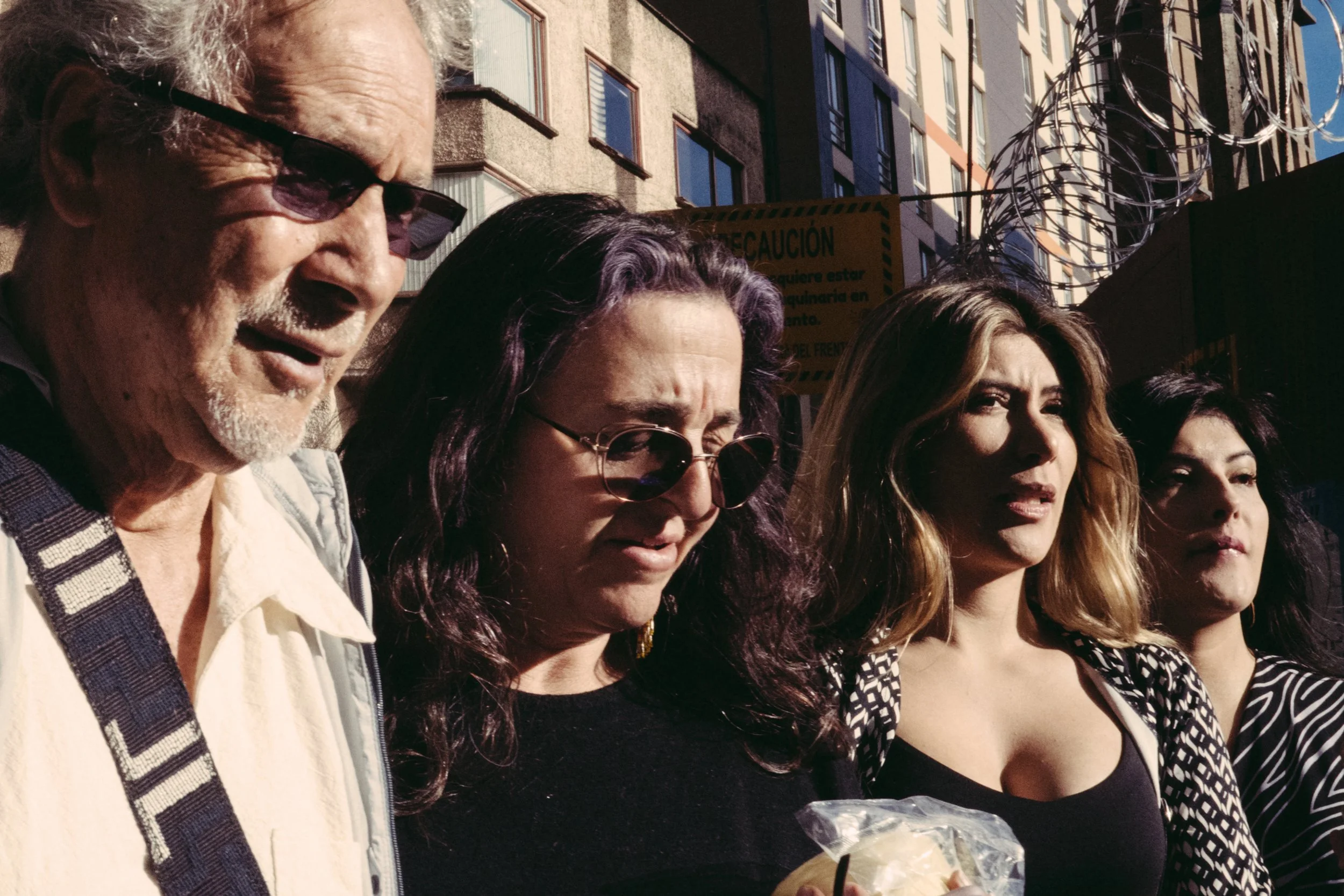 Grupo de personas en una fila, con fondo de edificios y barandales de alambre, algunas con gafas de sol, en un día soleado.