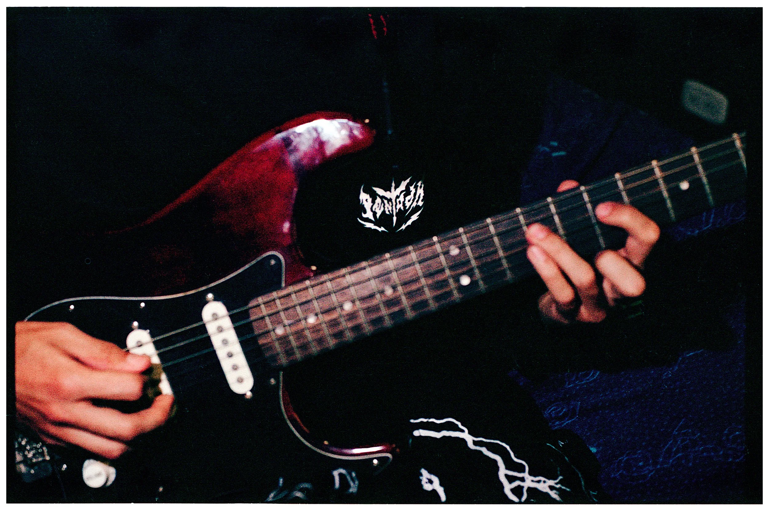 Persona tocando una guitarra eléctrica en un concierto o práctica, con manos en las cuerdas y el cuerpo de la guitarra visible.