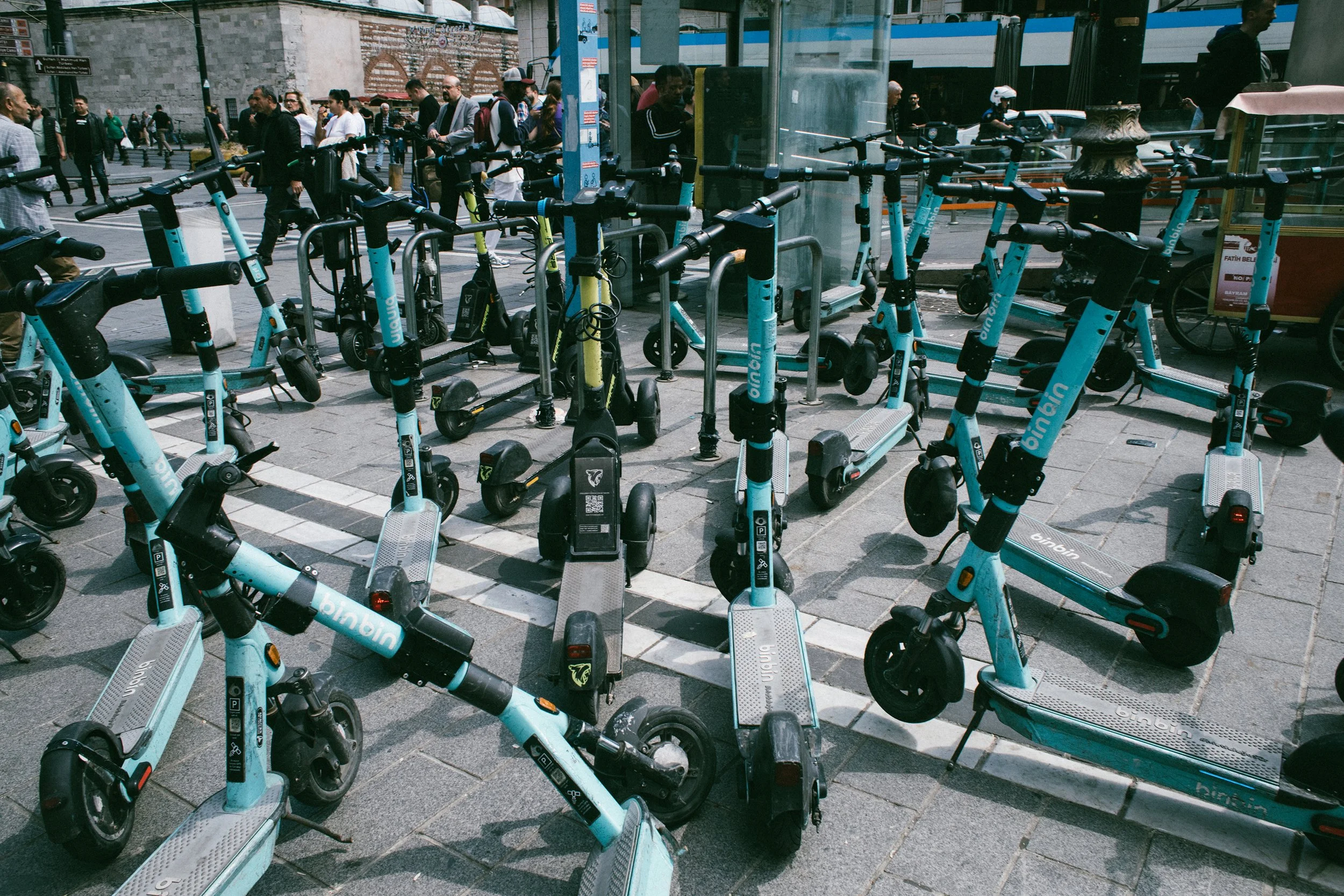 Una fila de patinetes eléctricos de color azul claros, algunos aparcados en un estacionamiento urbano, con varias personas caminando al fondo en una calle concurrida.