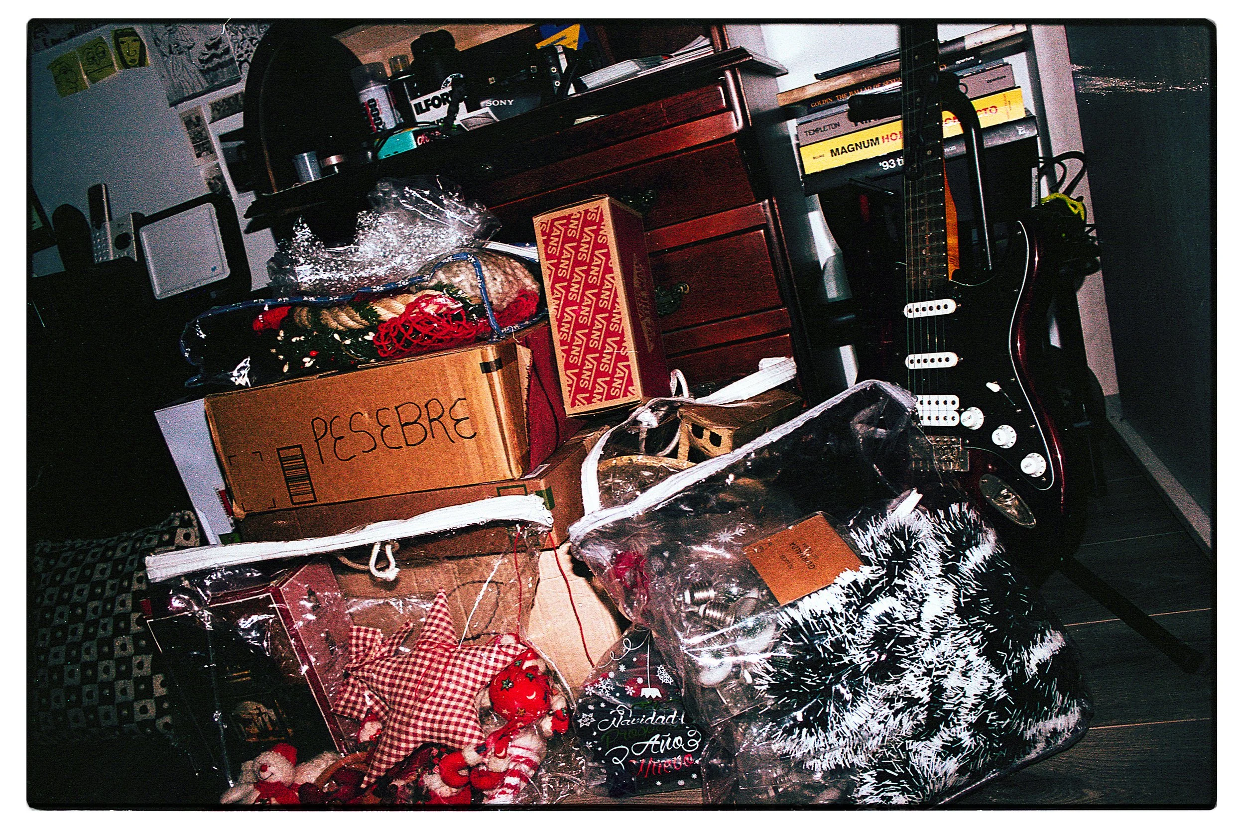 Mesa desordenada con regalos de navidad, bolsas decoradas, una guitarra eléctrica negra, libros, cajas de cartón, y decoraciones festivas.