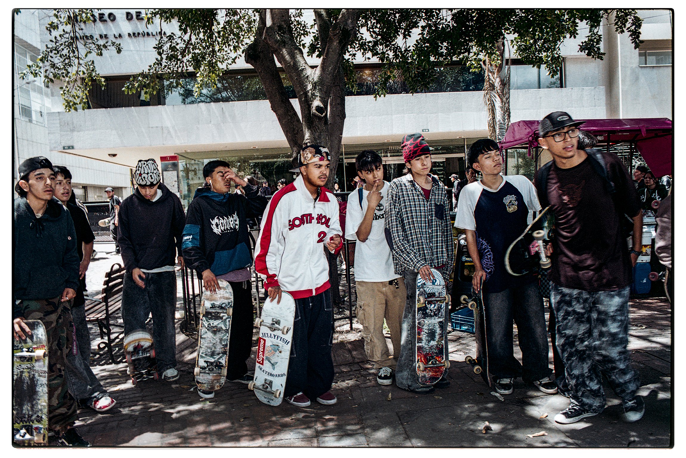 Grupo de jóvenes en la calle con patinetas, algunos llevan cascos y mochilas, algunos están apoyados en bancos, con árboles y edificios modernos en el fondo.
