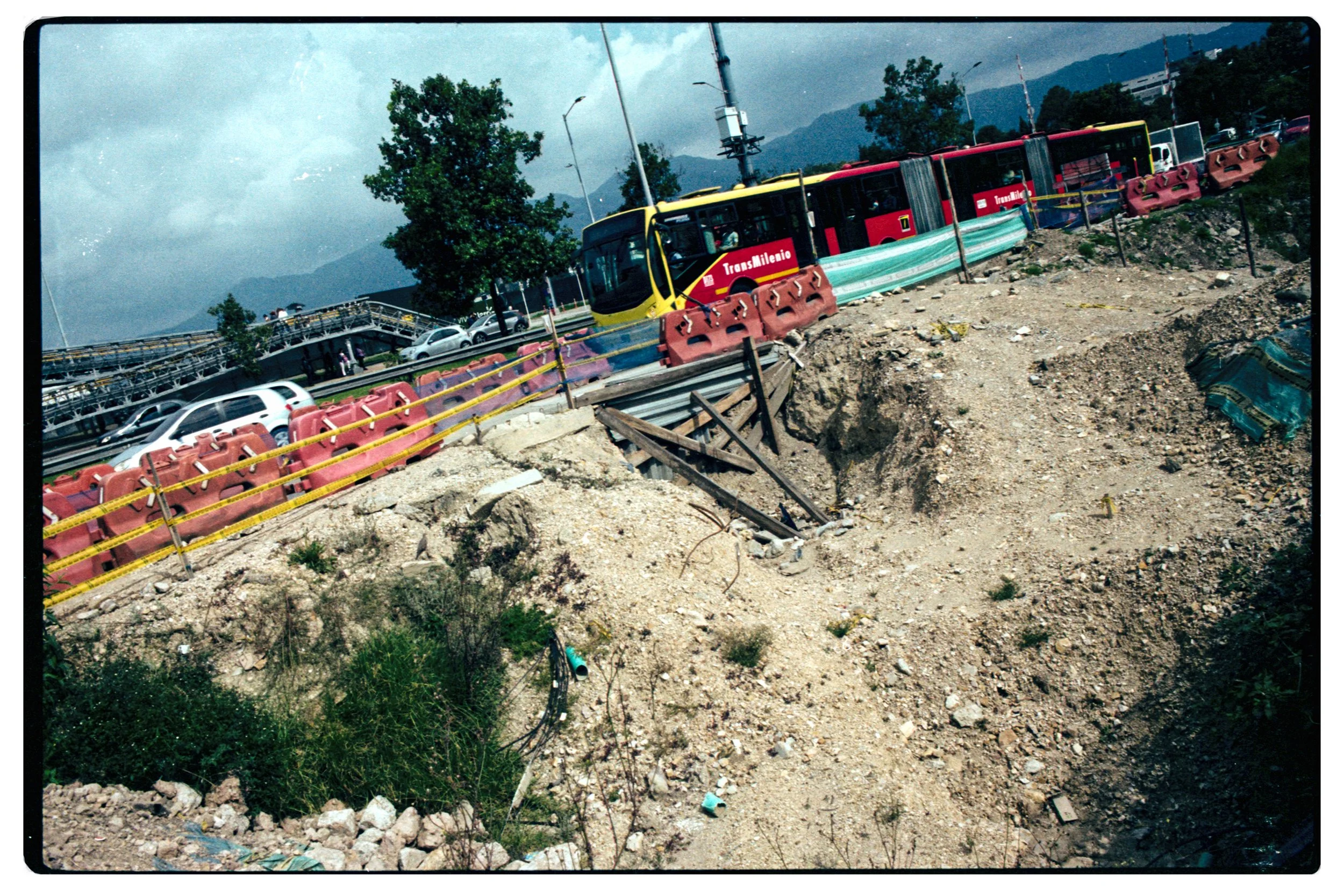 Una tramvía con pasajeros pasa por una zona en construcción con barreras de seguridad y tierra expuesta, al fondo hay árboles y montañas.