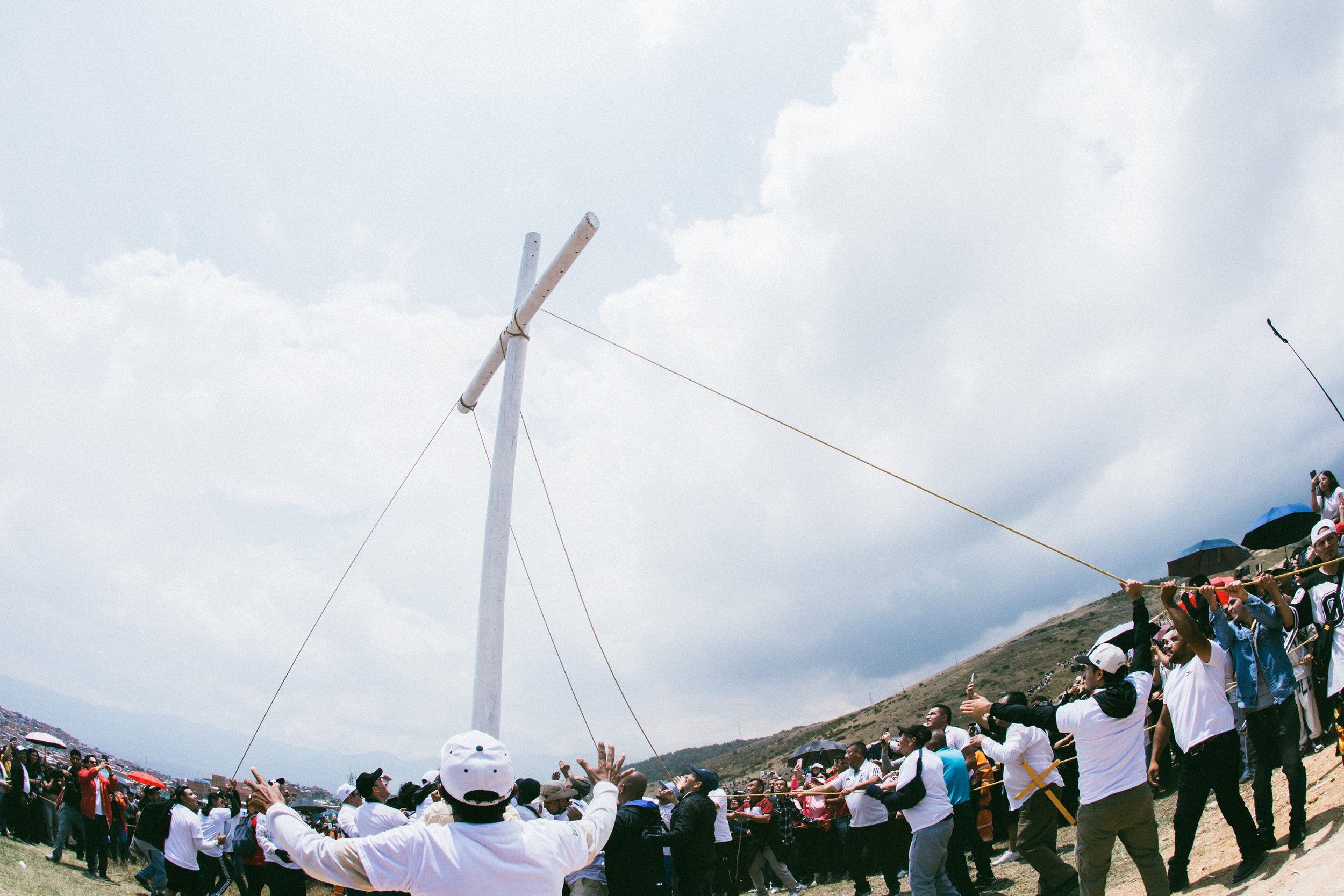 Un grupo de personas participando en una ceremonia cívica en un lugar al aire libre. Están sosteniendo una cuerda que se extiende hacia un poste, en un día nublado. Hay personas con sombreros y ropa casual, y en el fondo una colina.