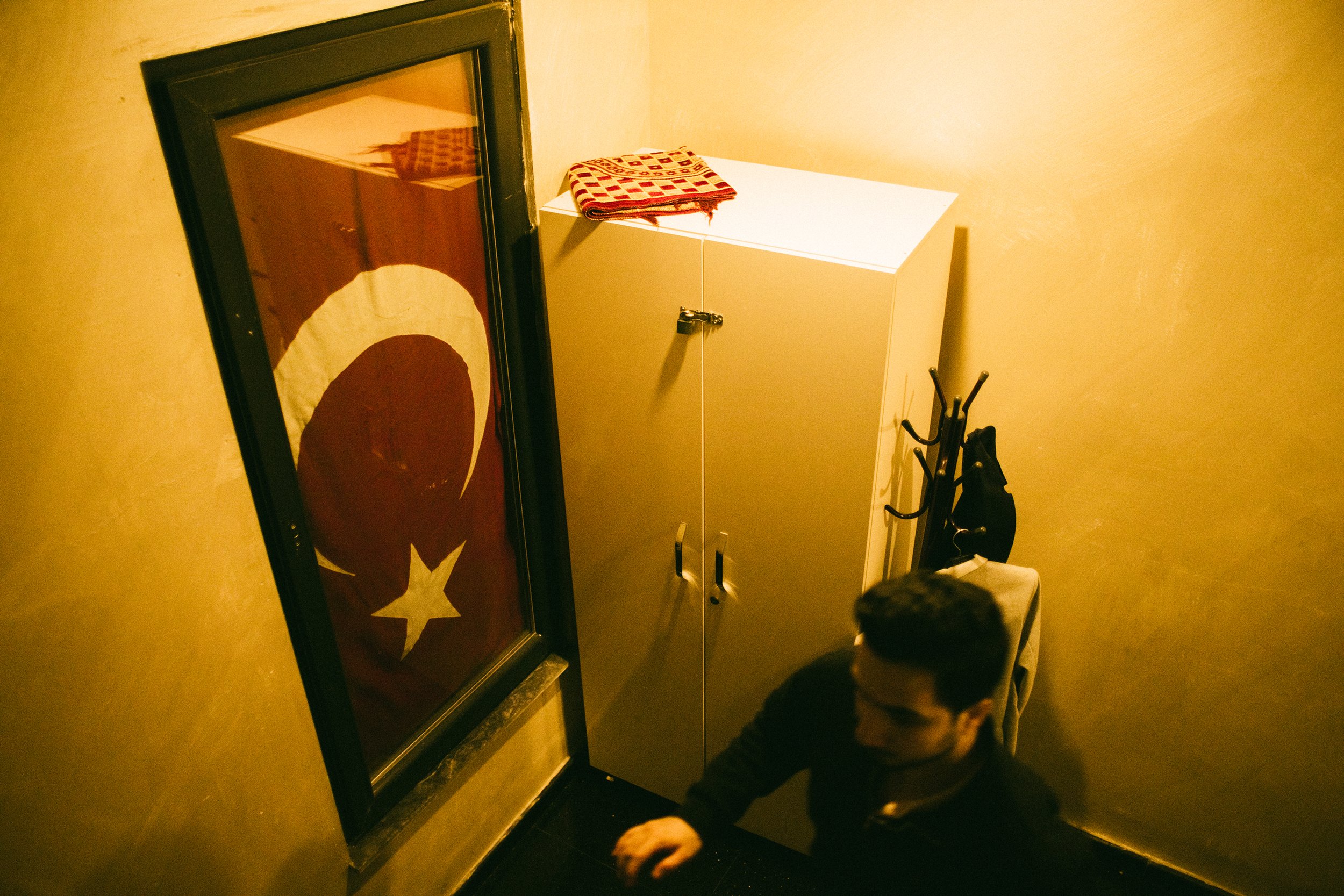 Una habitación con puertas de armario blancas en la esquina, sobre ellas una toalla con cuadros en tonos rojos y beige. A la izquierda, un cuadro con la bandera de Turquía en la pared y un espejo. En la esquina derecha, una persona de cabello oscuro 