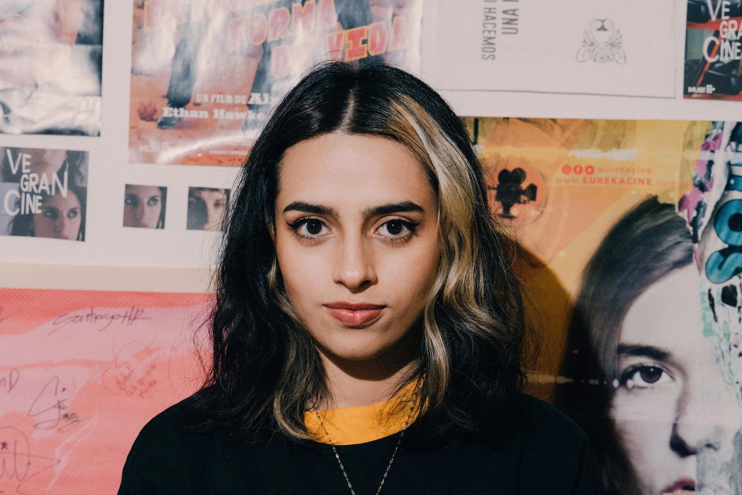 Retrato de una joven con cabello castaño y rubio, ojos grandes y cejas definidas, vestida con una camiseta color mostaza y suéter negro, posando frente a un mural con posters y fotografías.