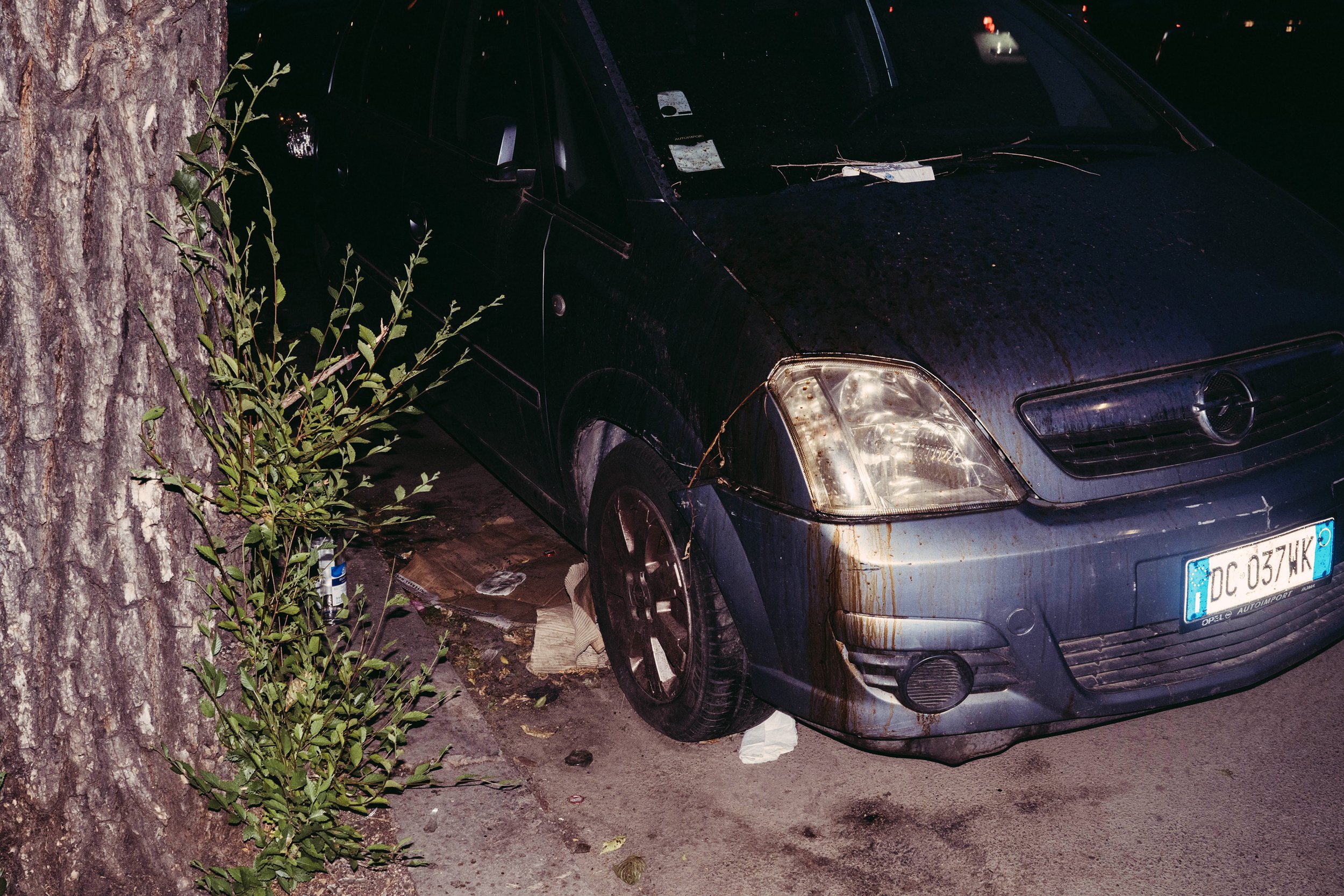 Coche negro estacionado junto a un árbol, con rastros de barro y objetos dispersos en el suelo, en una escena nocturna.