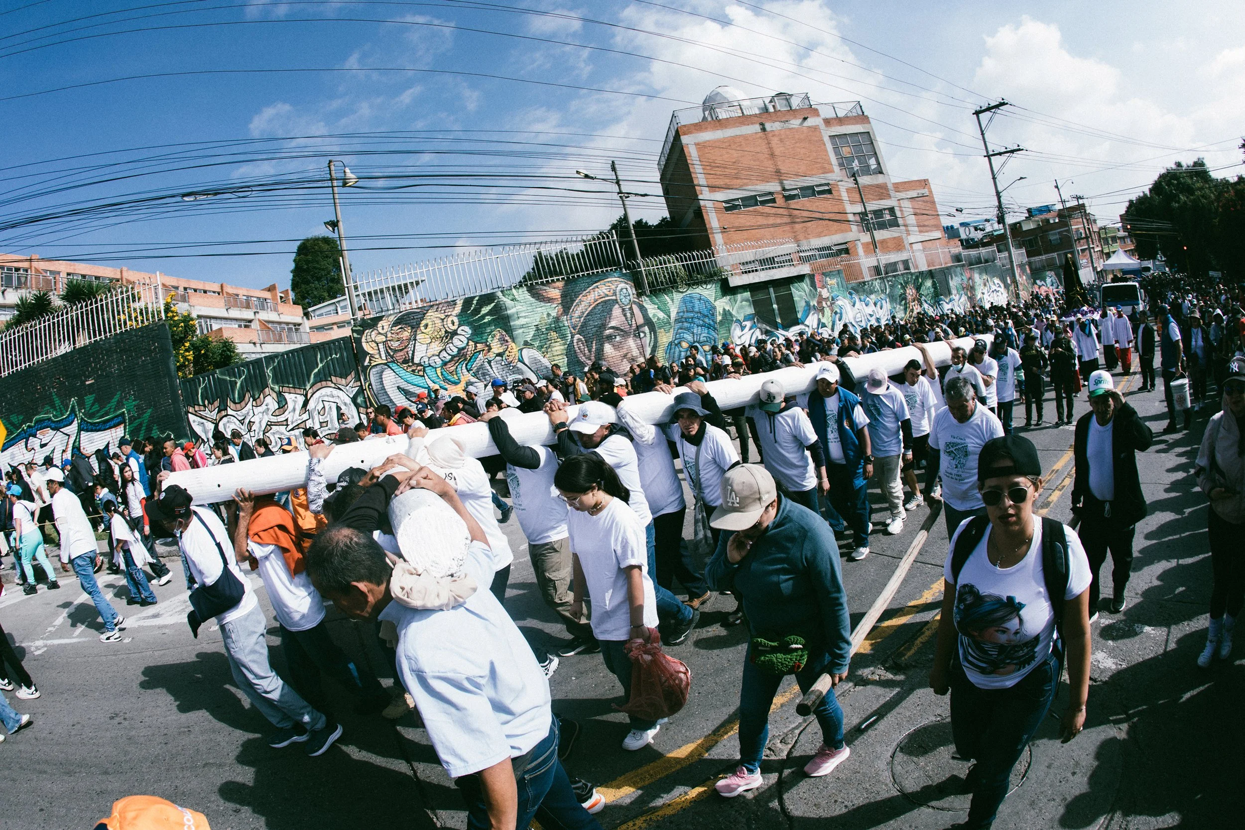 Personas participando en una marcha o manifestación en la calle, con una malla con arte urbano en la cerca y edificios al fondo bajo un cielo despejado.