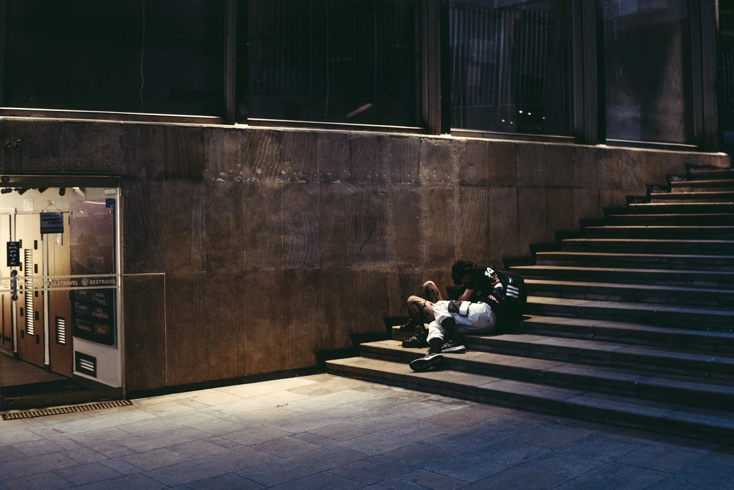 Persona descansando en la parte inferior de unas escaleras en un espacio interior con paredes de piedra. La escena está en penumbra con poca iluminación.