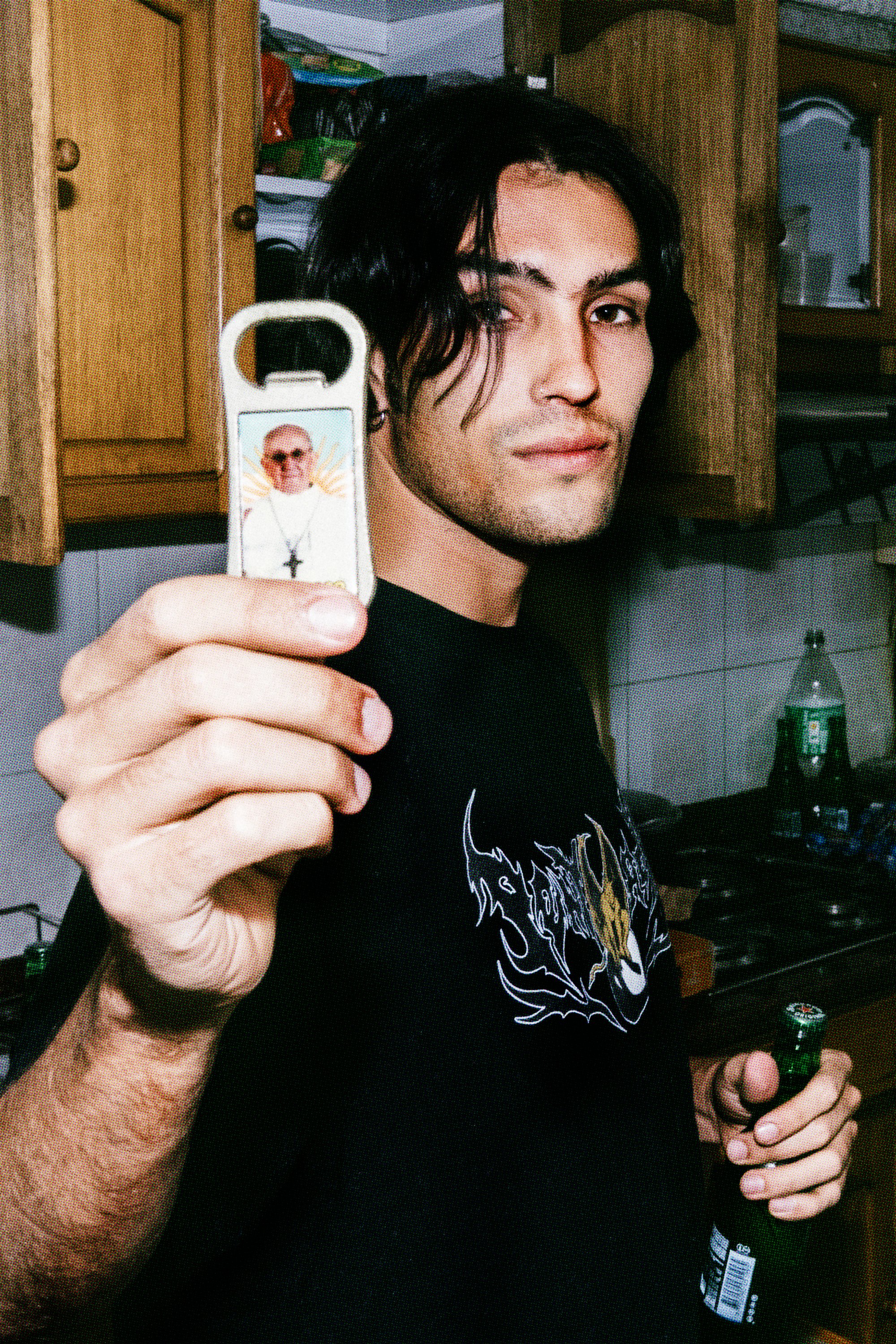 Joven con cabello negro y barba ligera sosteniendo una botella de cerveza, en un entorno de cocina con armarios de madera y varios objetos en el fondo.