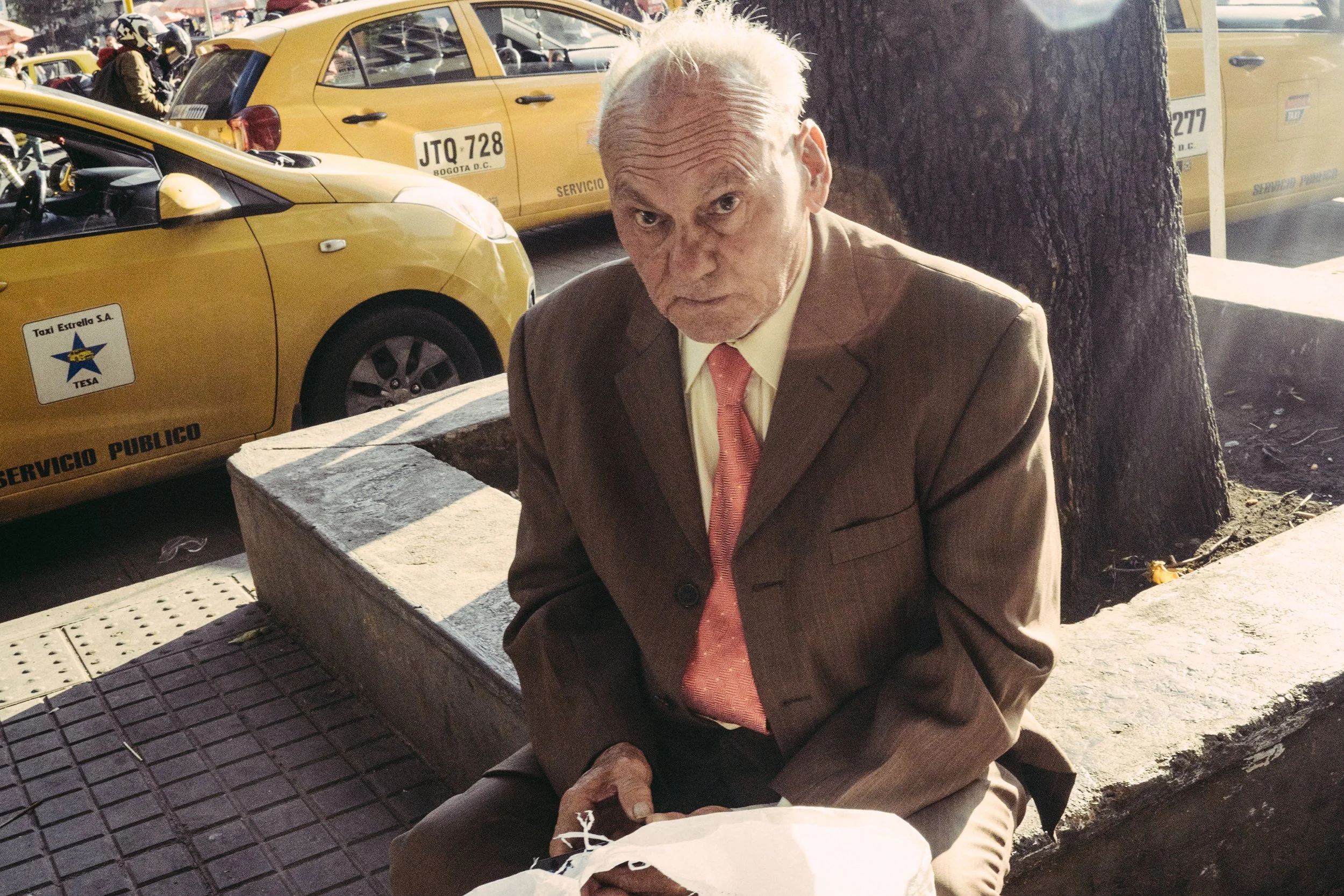 Hombre de cabello canoso con expresión severa, vestido con chaqueta marrón, camisa amarilla y corbata rosada, sentado en un banco en la acera, rodeado de taxis amarillos en una ciudad.
