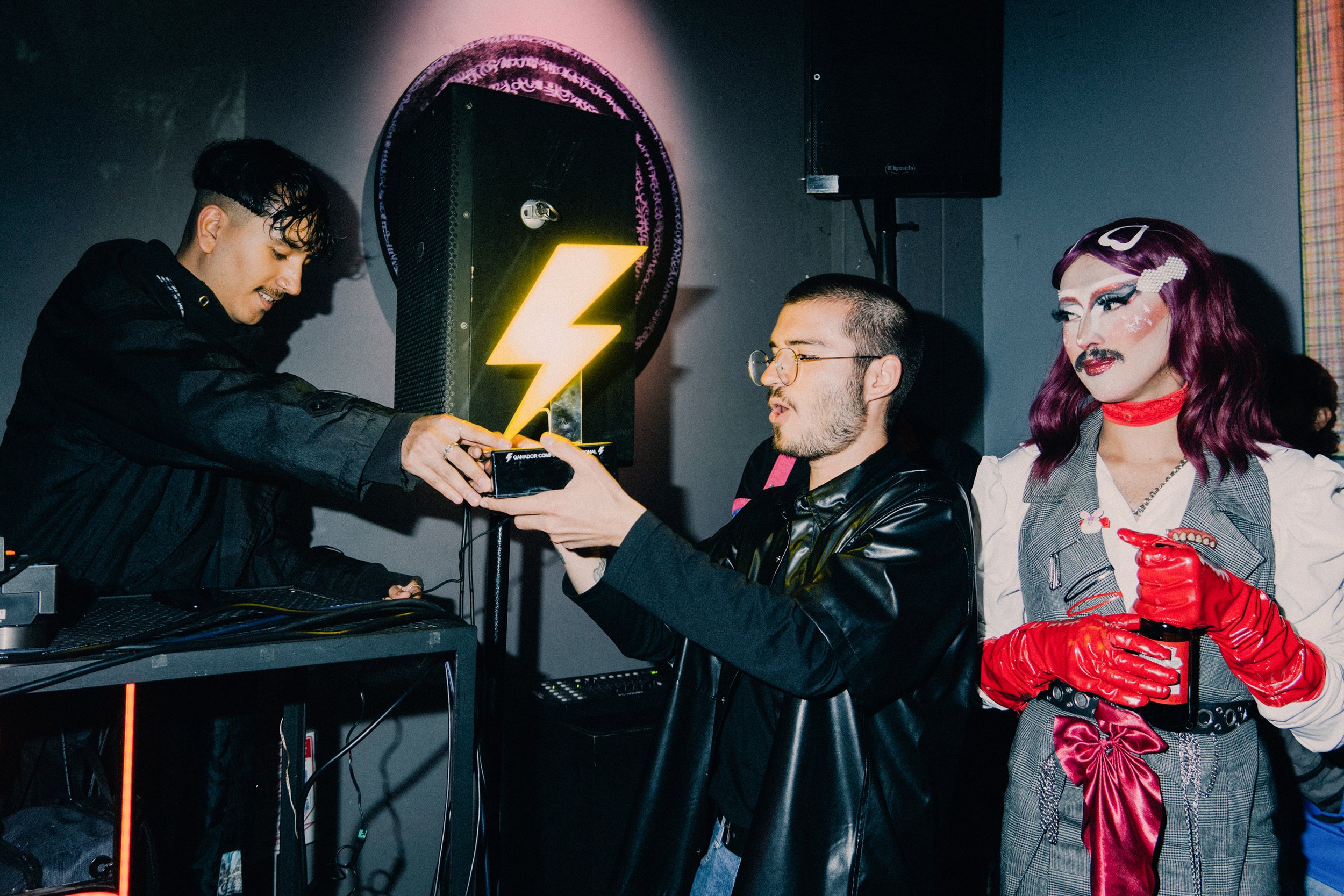Tres personas en un evento, una entregando un trofeo a otra, con una figura de rayo luminosa en el fondo y un grupo de personas en un entorno interior.