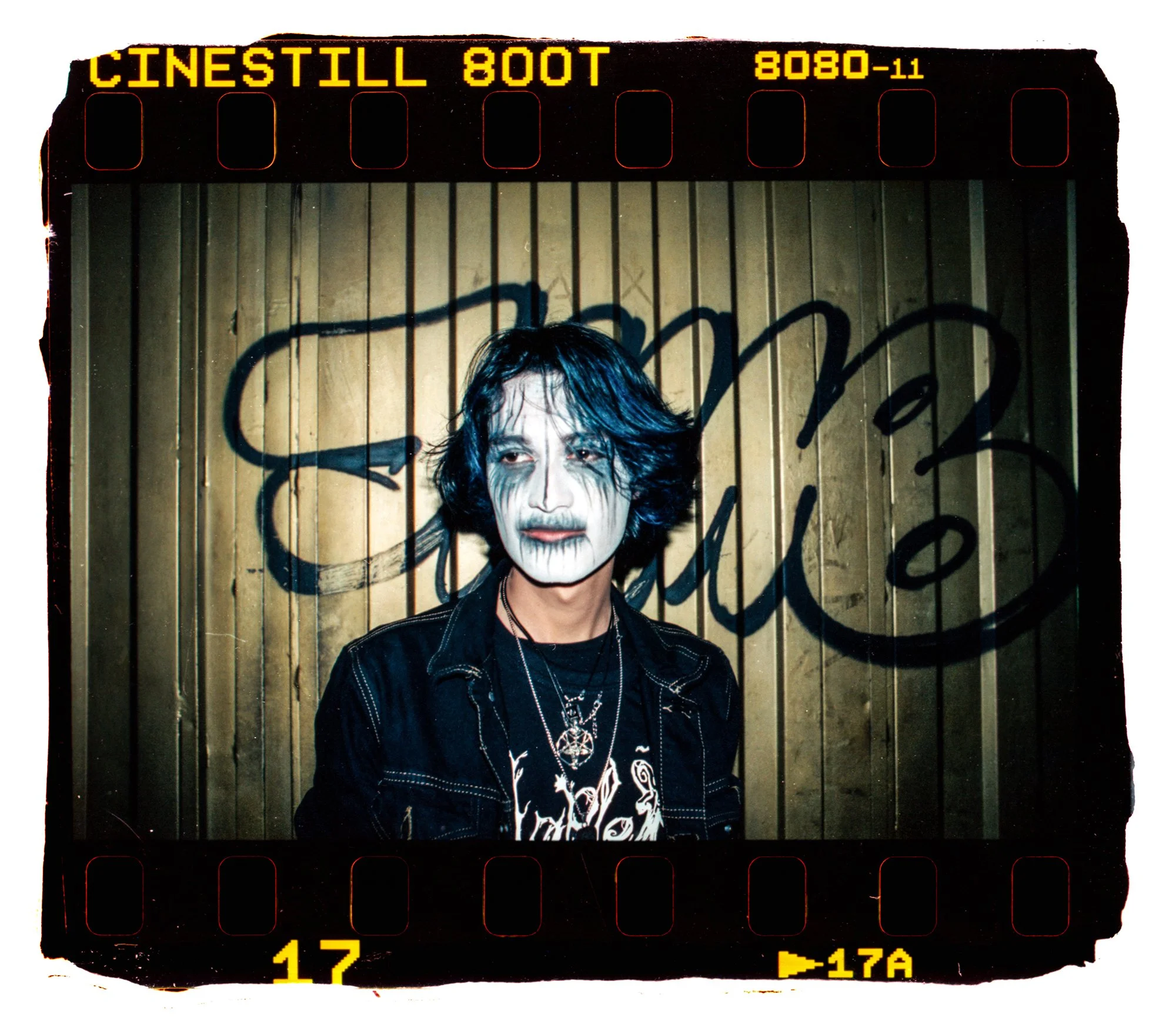 Retrato de un hombre con maquillaje de payaso, pelo largo y oscuro, vistiendo una chaqueta negra con un diseño de gráficos blancos y collares, frente a una puerta metálica decorada con graffiti y letras negras.
