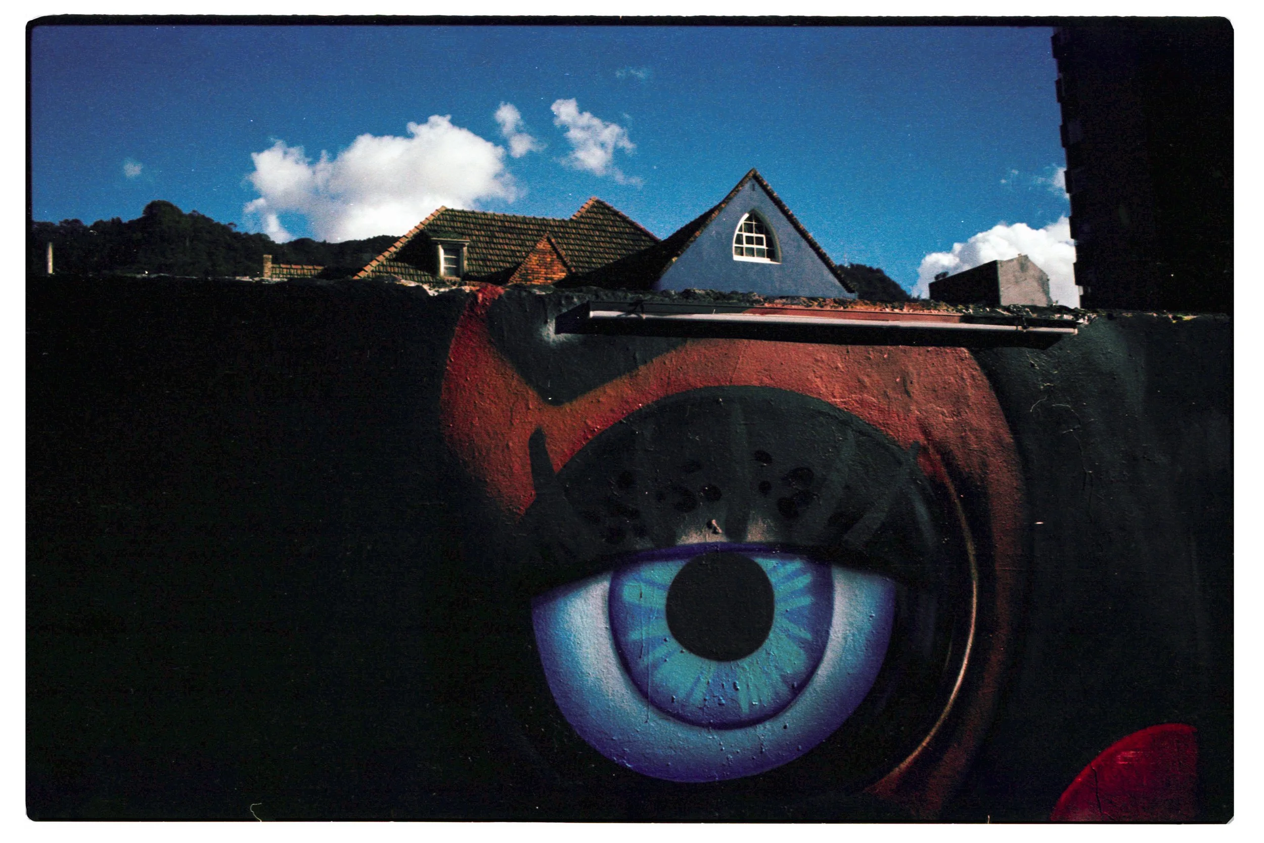Vista desde abajo de una casa con techo de tejas y ventanas, con un cielo azul y nubes blancas, y un mural de un ojo en la pared negra en primer plano.