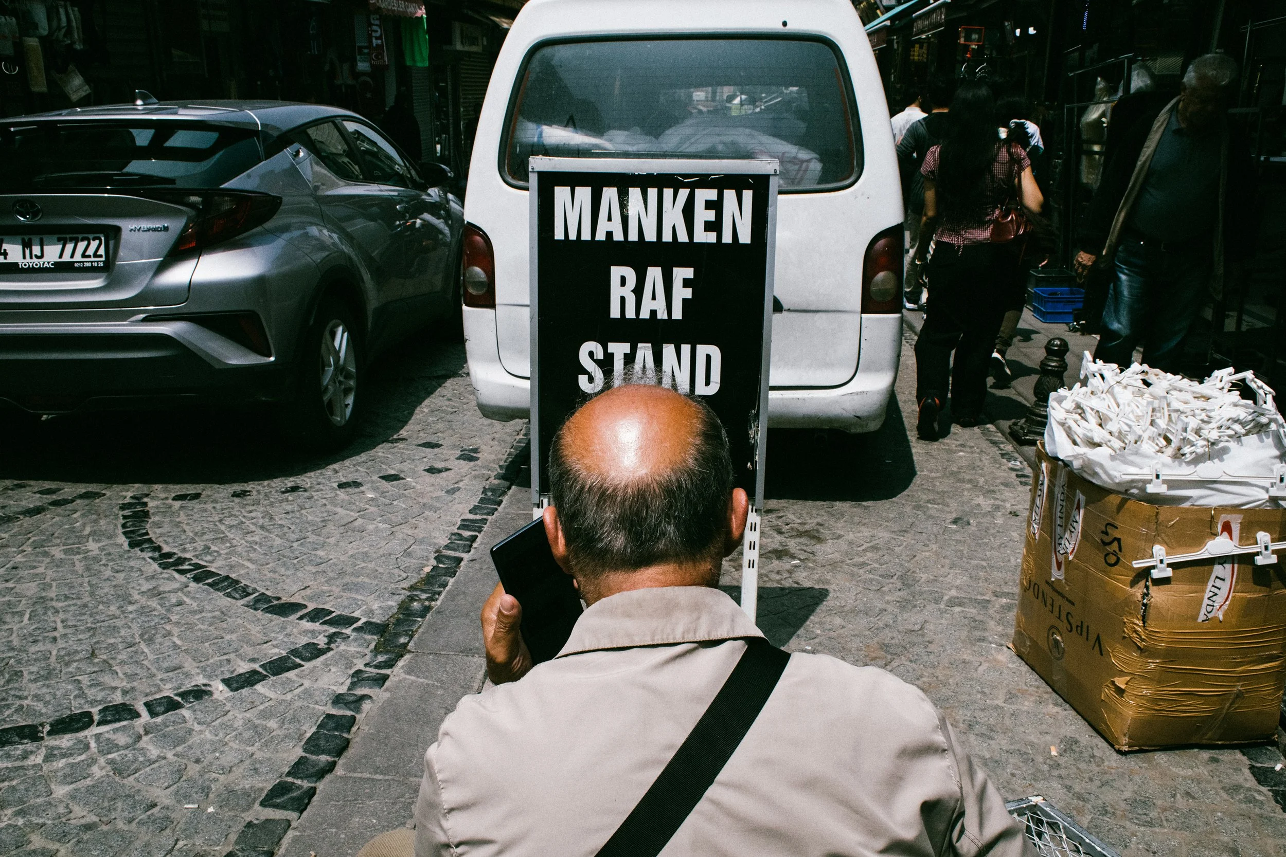 Persona con cabello canoso y camiseta beige, sentado en la calle, sosteniendo un teléfono, frente a un cartel que dice 'MANKEN RAF STAND'. Detrás hay autos, personas caminando y un puesto con pequeños objetos y bolsas de plástico blancas.