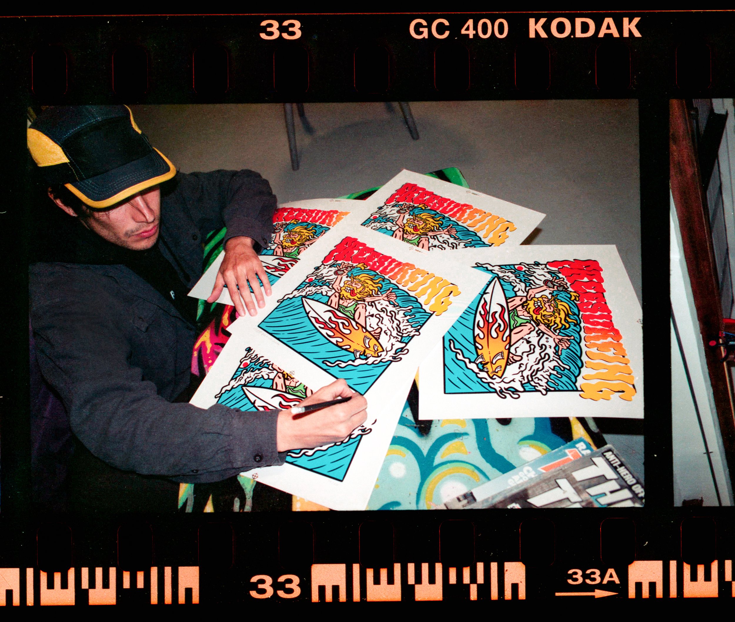 Un hombre con gorra negra y amarilla está firmando posters de colores vivos con una ilustración de un diablo en un escritorio, debajo de una barra con texto y marcas de medición.