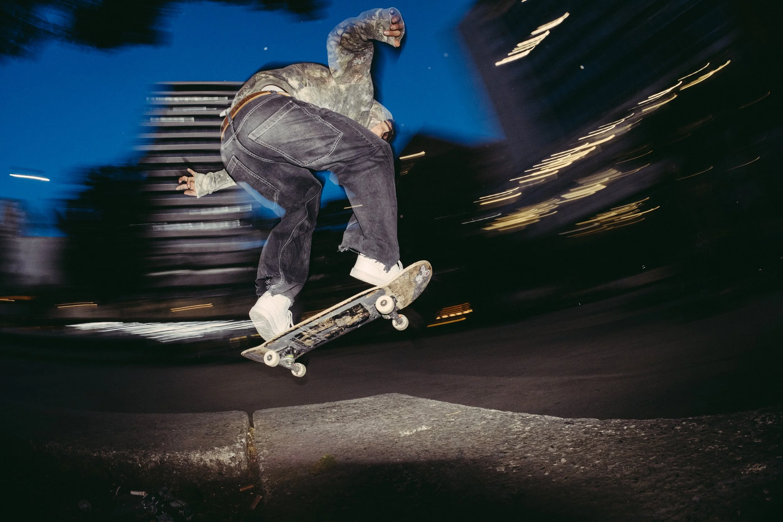 Un joven haciendo trucos de skate en la calle por la noche, con luces borrosas en el fondo.