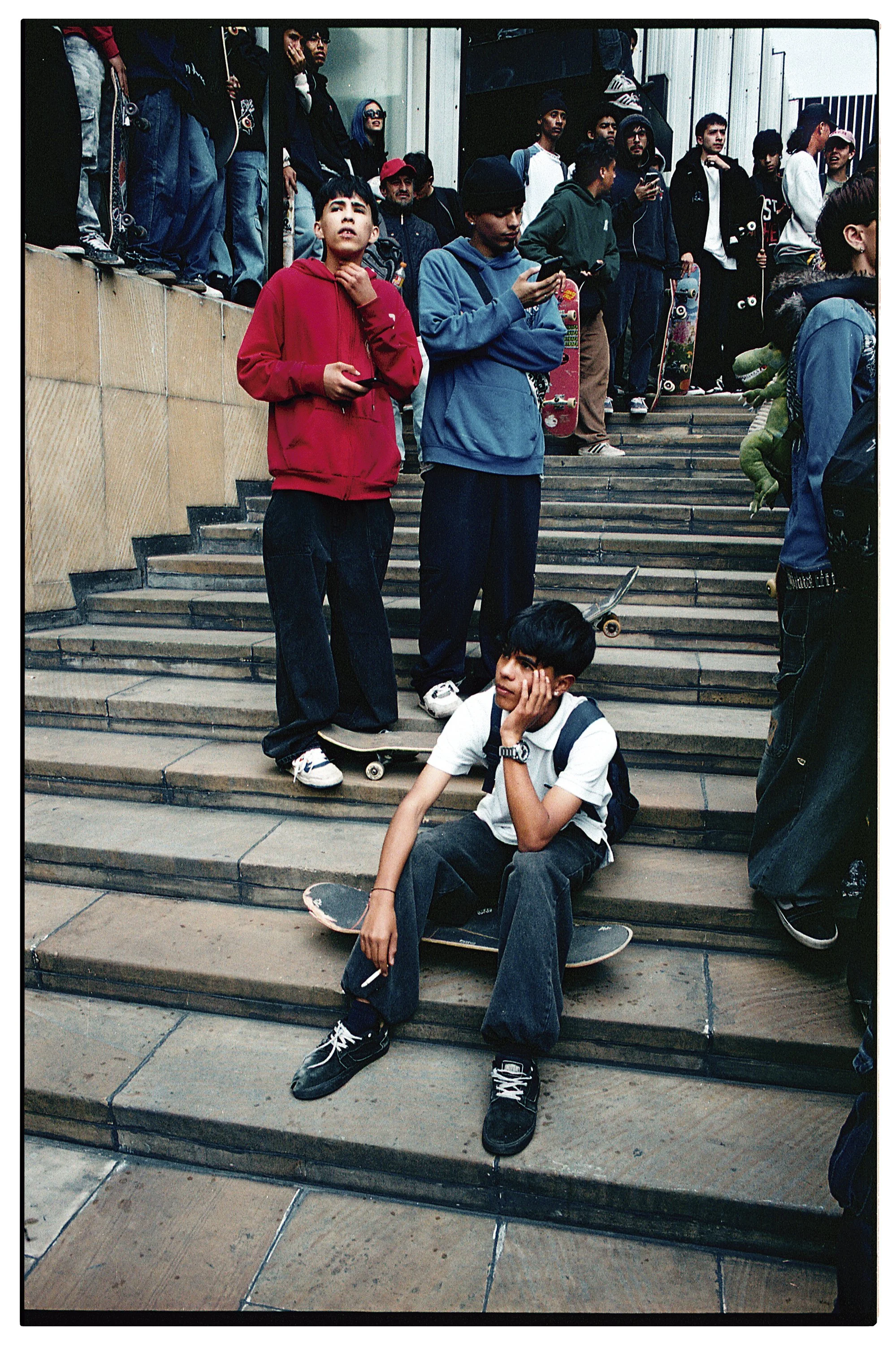 Grupo de jóvenes en las escaleras, algunos con patineta, algunos usando teléfonos móviles, algunos con skateboards, en un entorno urbano.