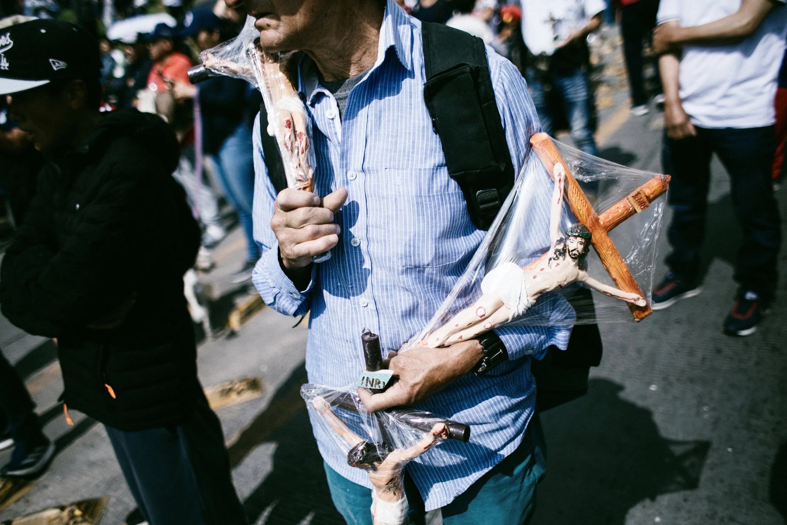 Hombre sosteniendo una cruz de madera con la imagen de Jesús, envuelta en plástico, en una marcha o manifestación con otras personas alrededor.