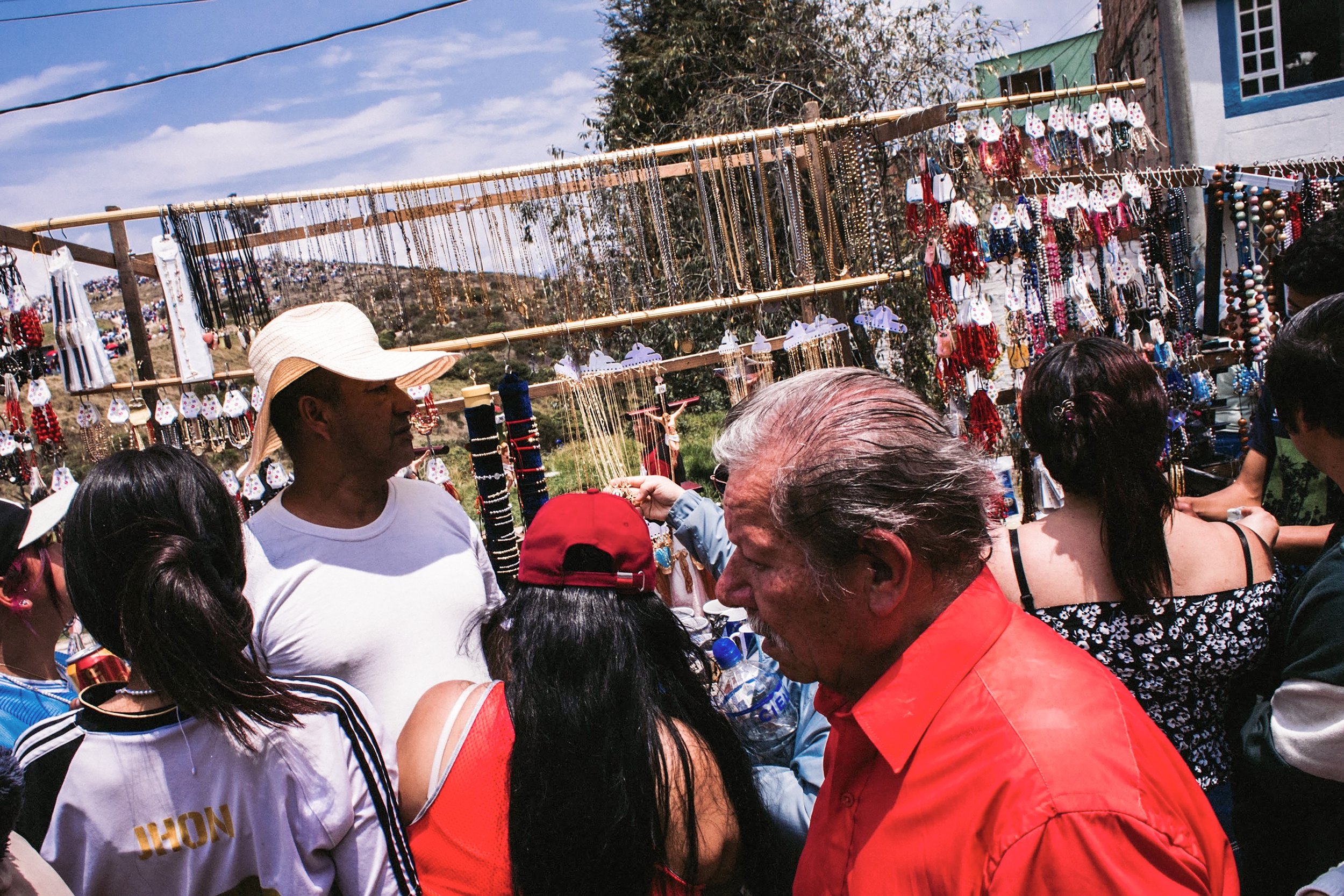 Personas en una feria con puestos de joyería y accesorios, en un día soleado y con cielo parcialmente nublado.