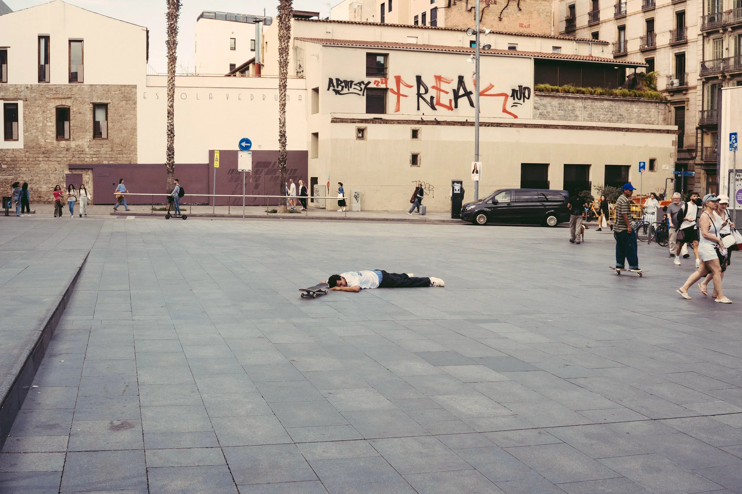 persona acostada en el suelo en un espacio público, con una patineta cerca, rodeada de otras personas caminando, con edificios y graffiti en la pared de fondo