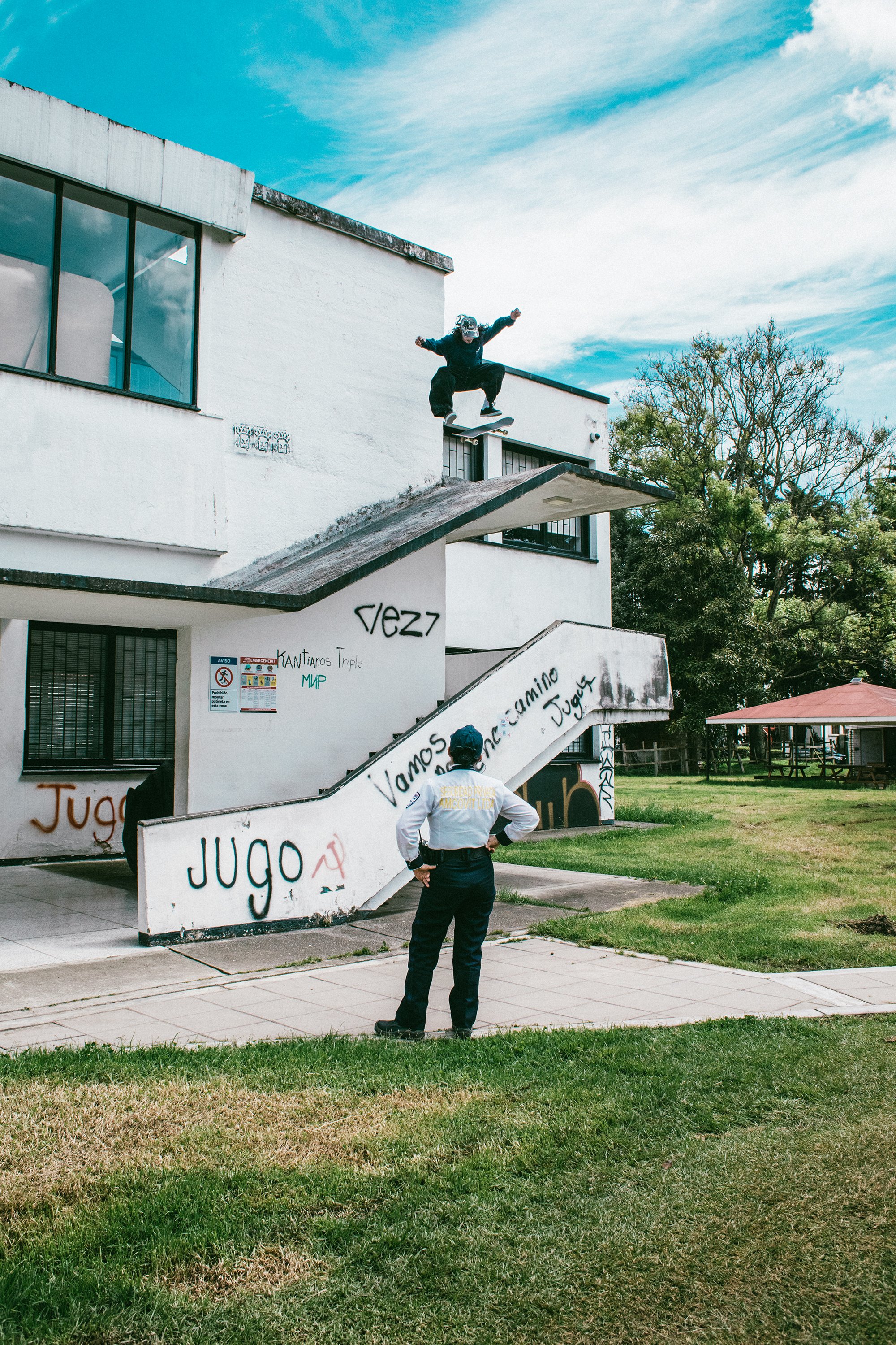 Persona observando a un skater realizando un truco en un borde de un edificio en un espacio abierto con césped y árboles.