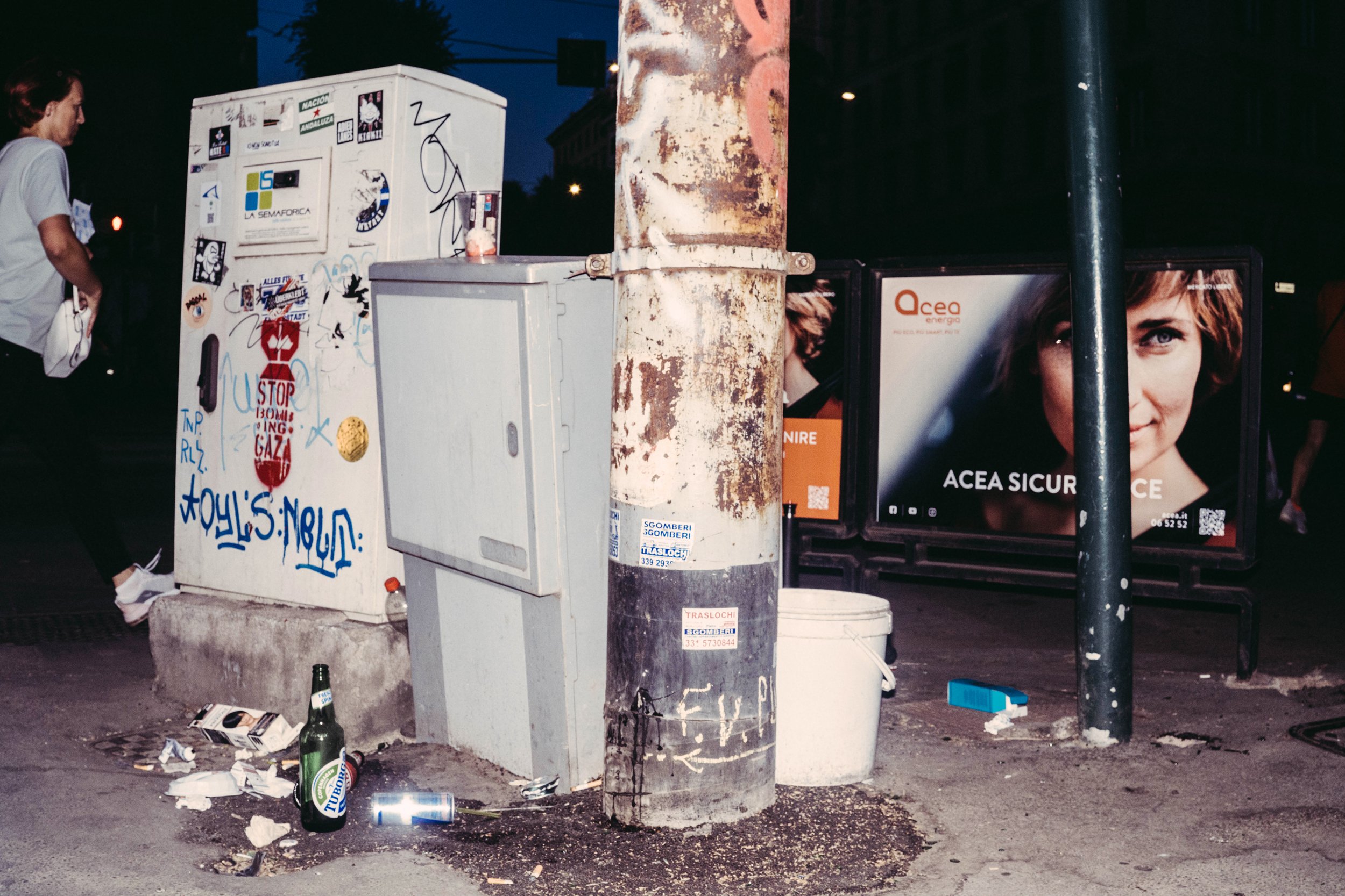 Escultura de una cara humana en el poste de una parada de autobús rodeada de basura y papel disperso en la calle por la noche.
