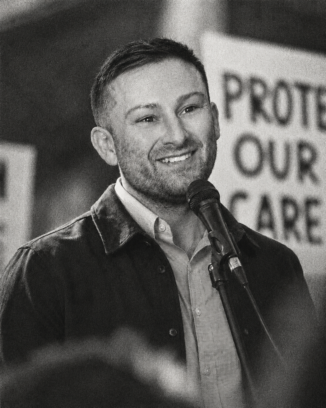 Aaron Welcher, smiling, standing at a microphone during a speech, in front of a sign that reads 'Protect Our Care'.