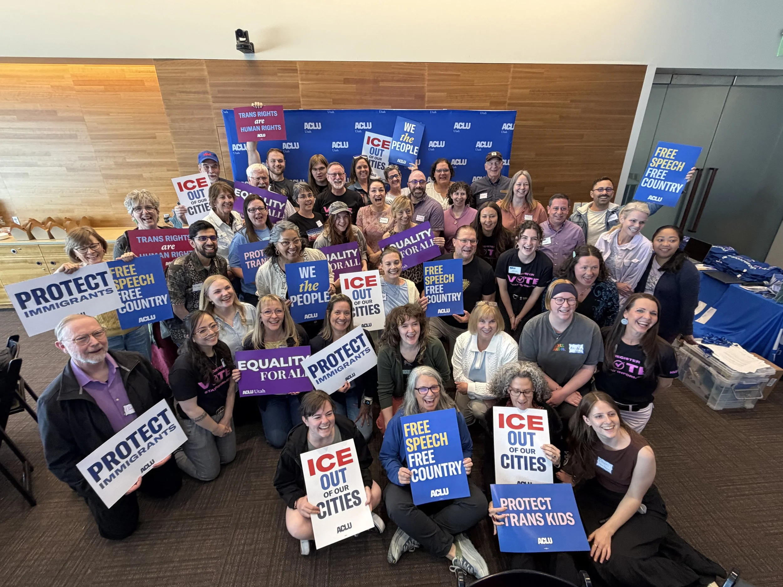 A diverse group of people at a rally holding signs with messages supporting immigrant rights, free speech, and trans rights, in front of a blue backdrop with the ACLU logo.