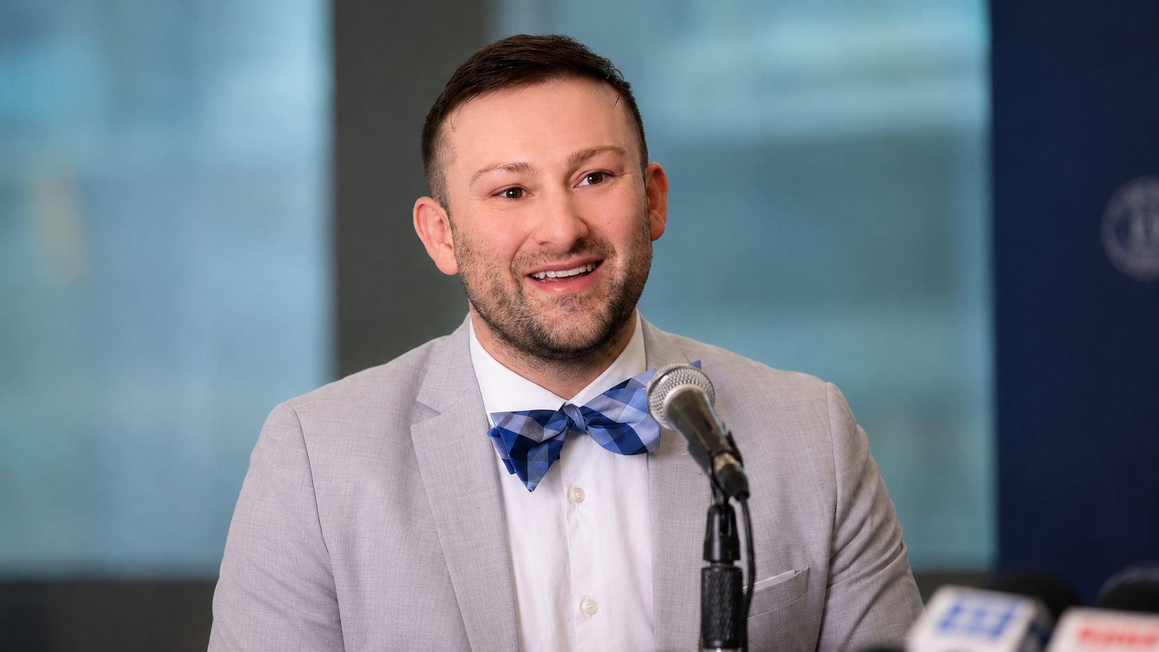 A smiling man in a light gray suit and blue checkered bowtie speaking at a press conference with microphones in front of him.