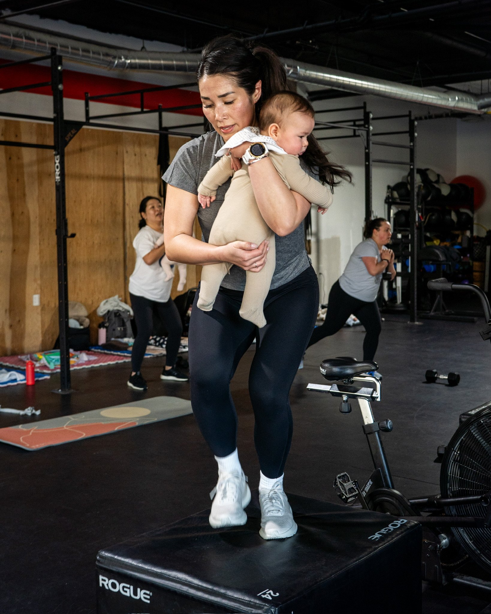 A woman holding a baby at a gym, surrounded by other women exercising on mats and fitness equipment.