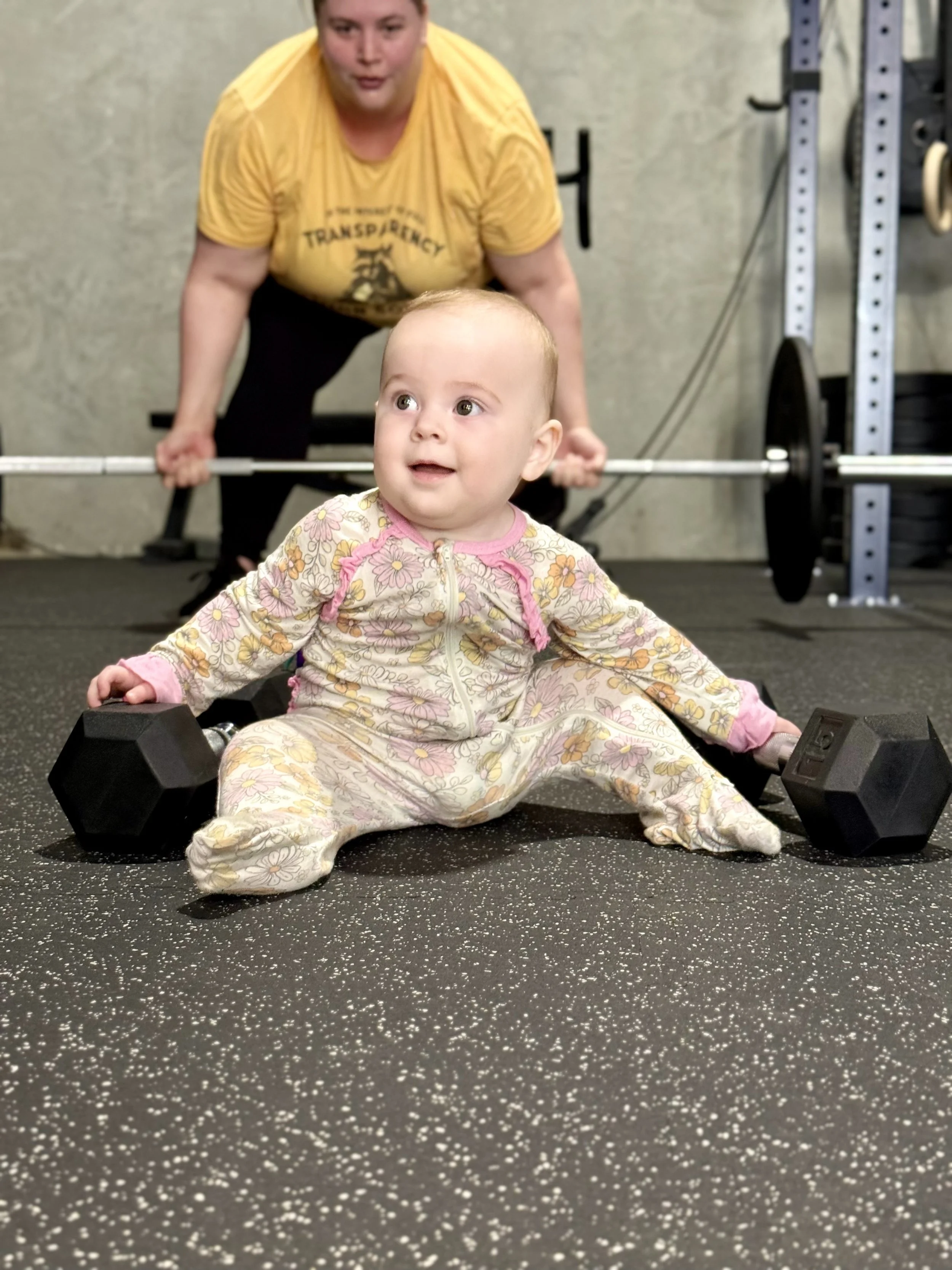A young child in floral pajamas sitting on the gym floor with dumbbells on either side, while a woman in a yellow shirt with the word "Transperency" watching in the background.