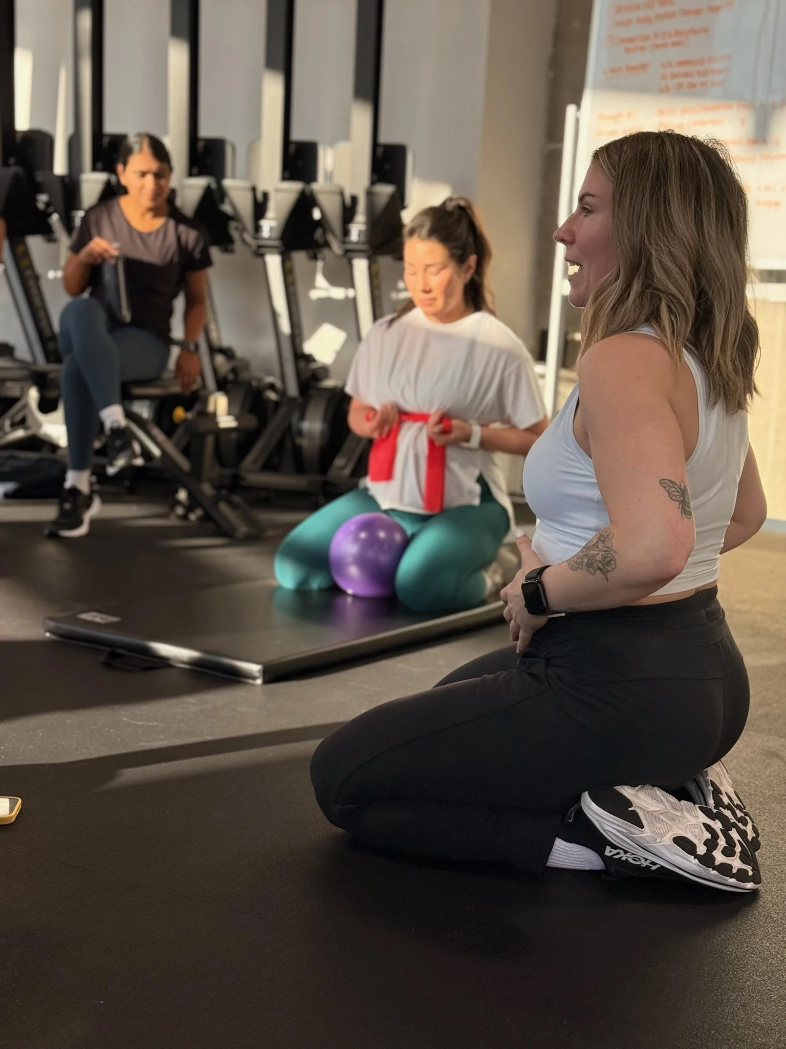 A woman with tattoos and blonde hair kneeling on the floor in a gym, facing to the right, while two women with darker hair and a woman with black hair are seated in front of her. The woman kneeling is wearing a white tank top, black pants, and athletic shoes. The woman in the middle is sitting cross-legged on a yoga mat, wearing a white shirt with a red belt, and appears to be using a fitness device. The woman on the left is seated with her legs crossed, and the woman in the background is standing next to exercise machines.