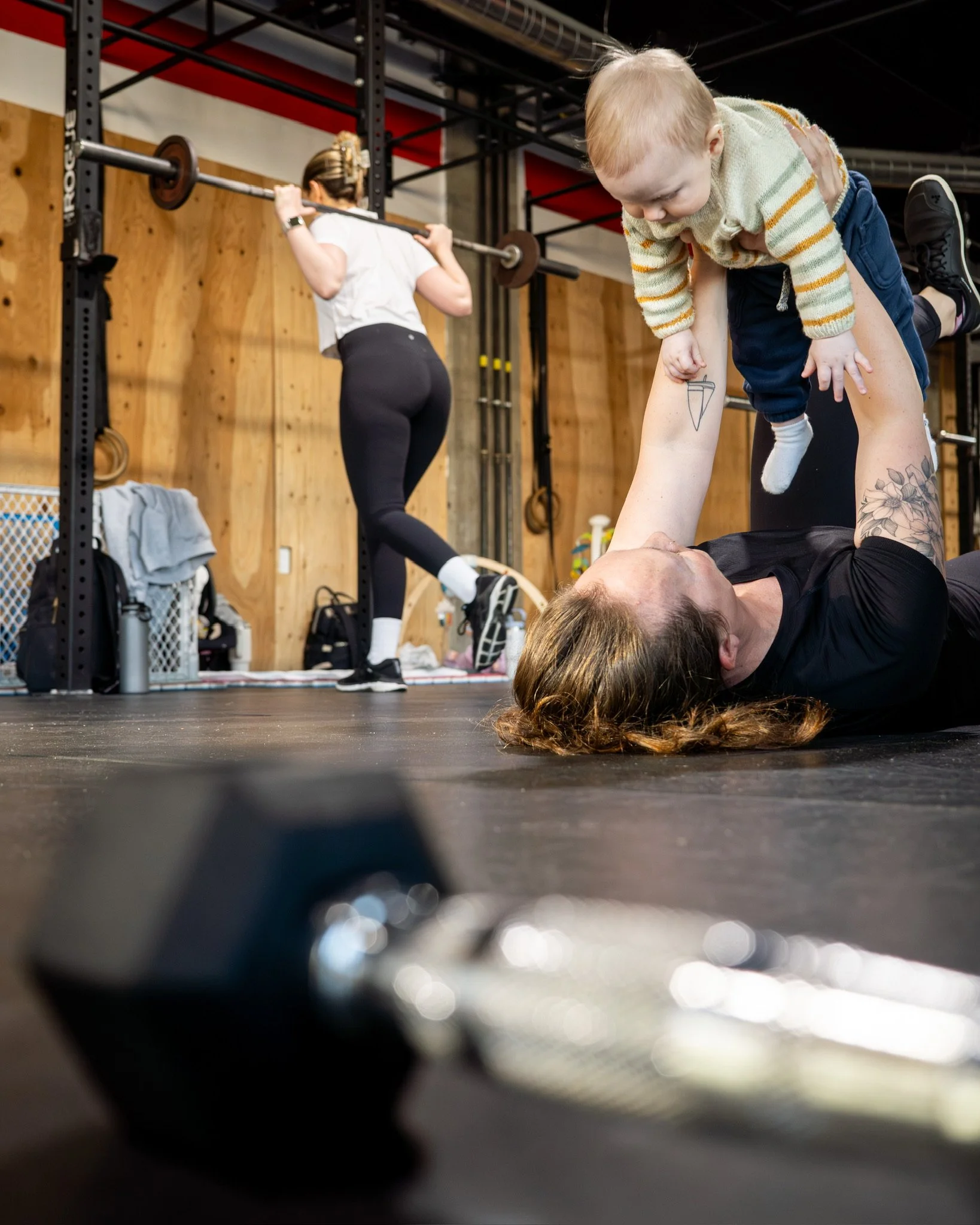 A child doing a handstand on the chest of a woman lying on the floor in a gym, with a woman lifting a barbell in the background.
