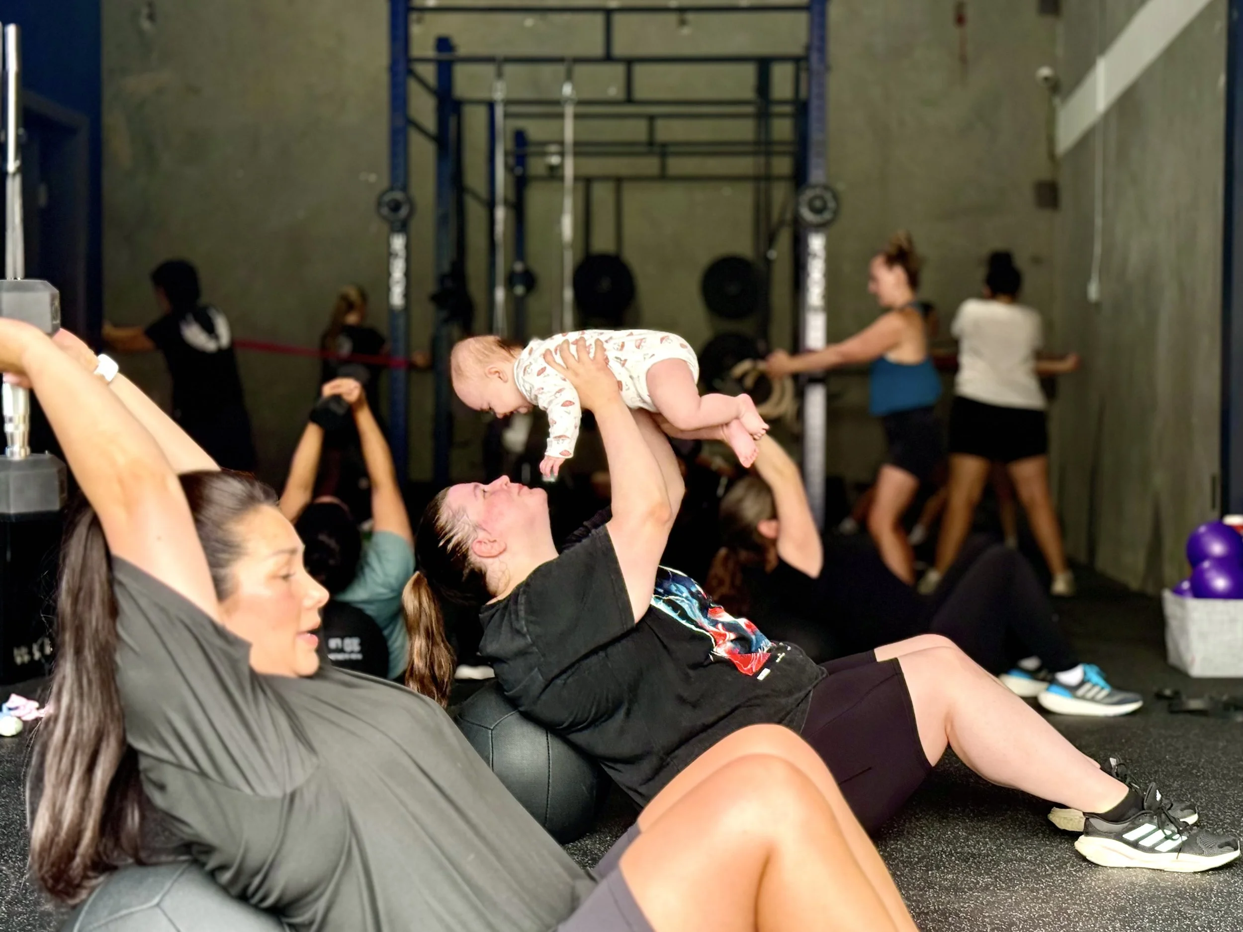 People exercising in a gym, with a woman holding a baby up in the foreground. Several others are doing exercises in the background near equipment.