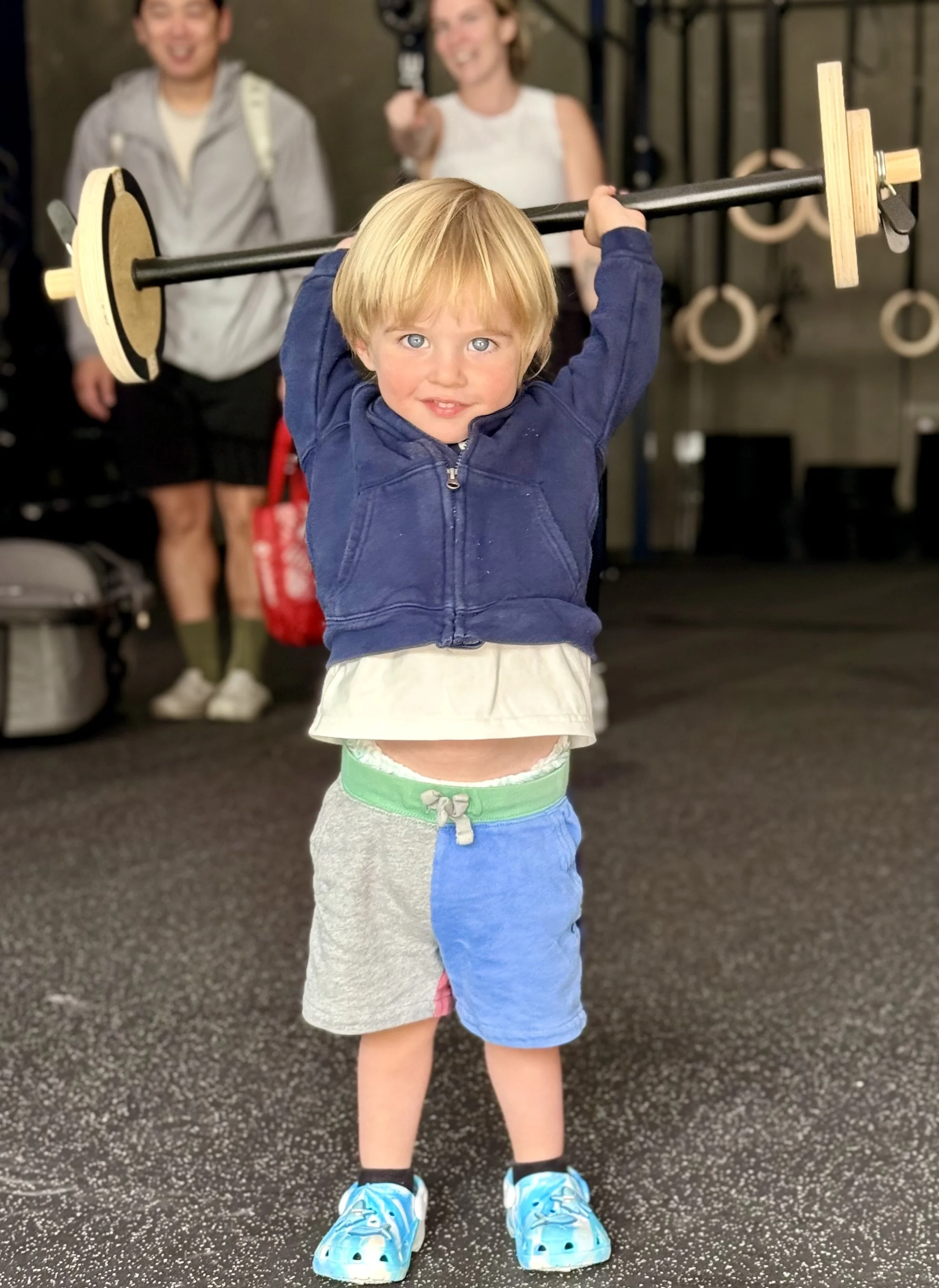 Child with blonde hair lifting a barbell in a gym, wearing a navy blue hoodie, gray shorts, and blue sneakers, with two smiling women in the background.