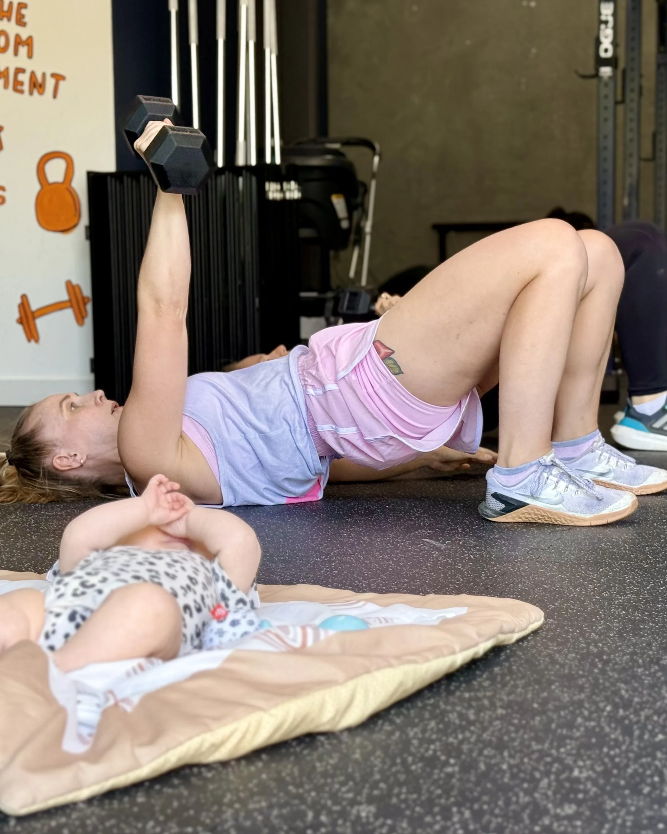 A woman lying on her side on a mat, lifting a small dumbbell with her arm, in a gym setting.