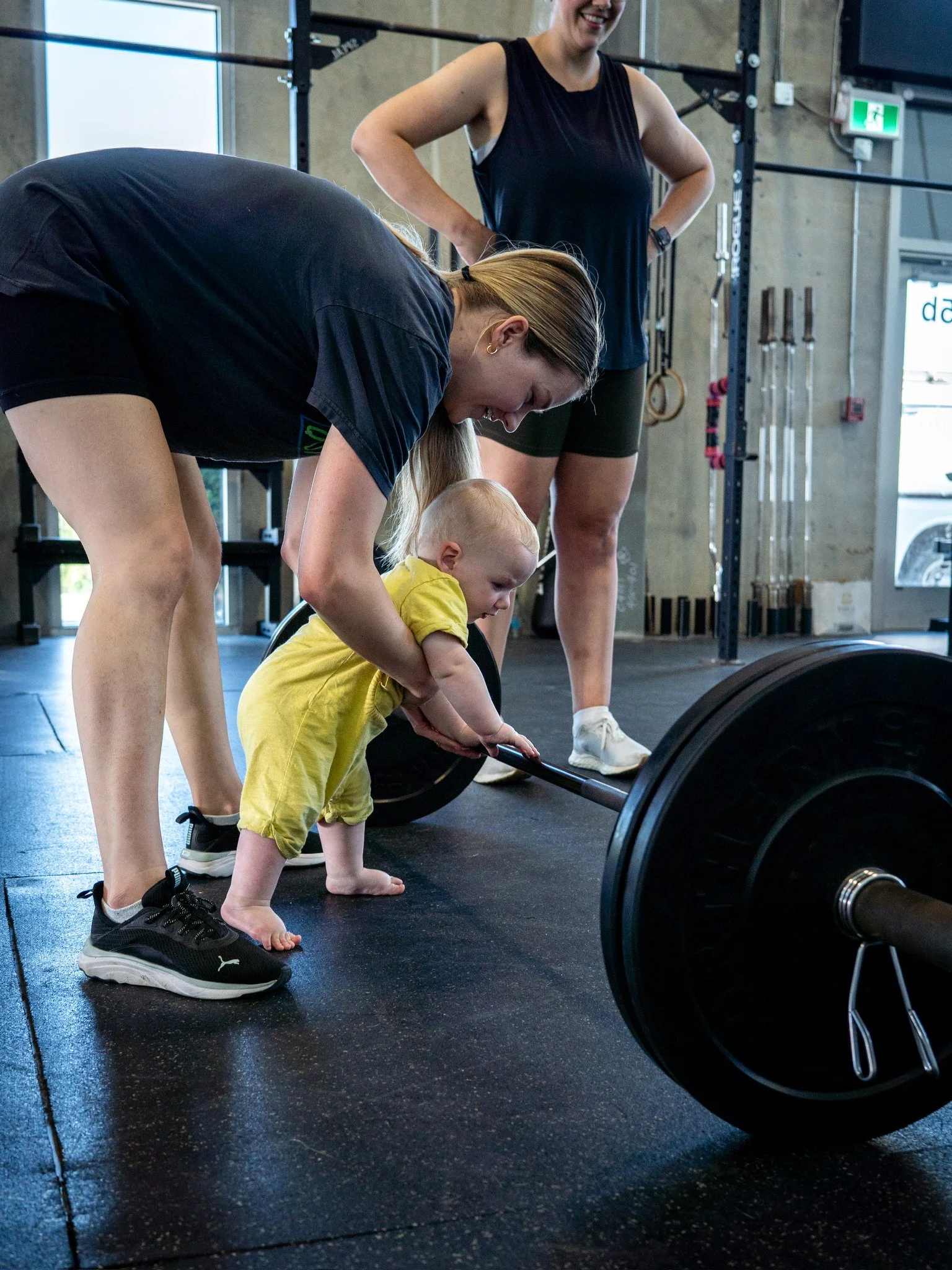 A young child with blonde hair and a yellow shirt, barefoot, holding a barbell with the help of a woman in a black shirt and shorts, in a gym with other women and gym equipment in the background.