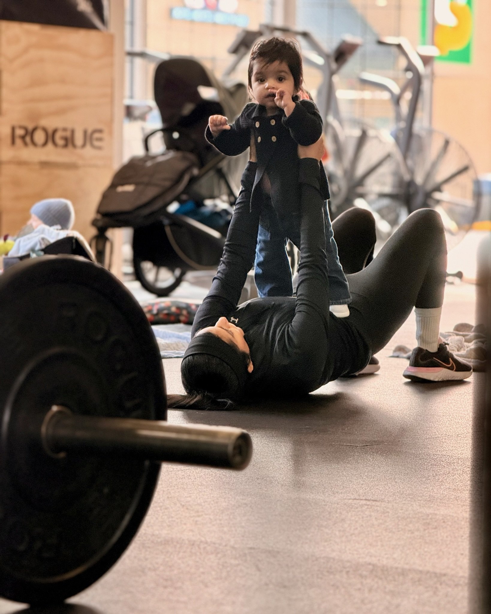 A woman lying on the floor lifting a young child in the air during playtime in a gym or workout area. The child has dark hair and is wearing a black jacket and jeans. Exercise equipment is visible in the background.