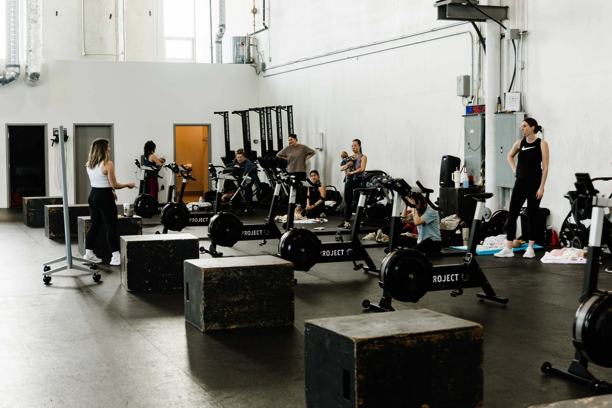 Indoor gym with women using exercise equipment, some sitting or standing, some holding babies, with busy atmosphere and fitness branding on equipment.