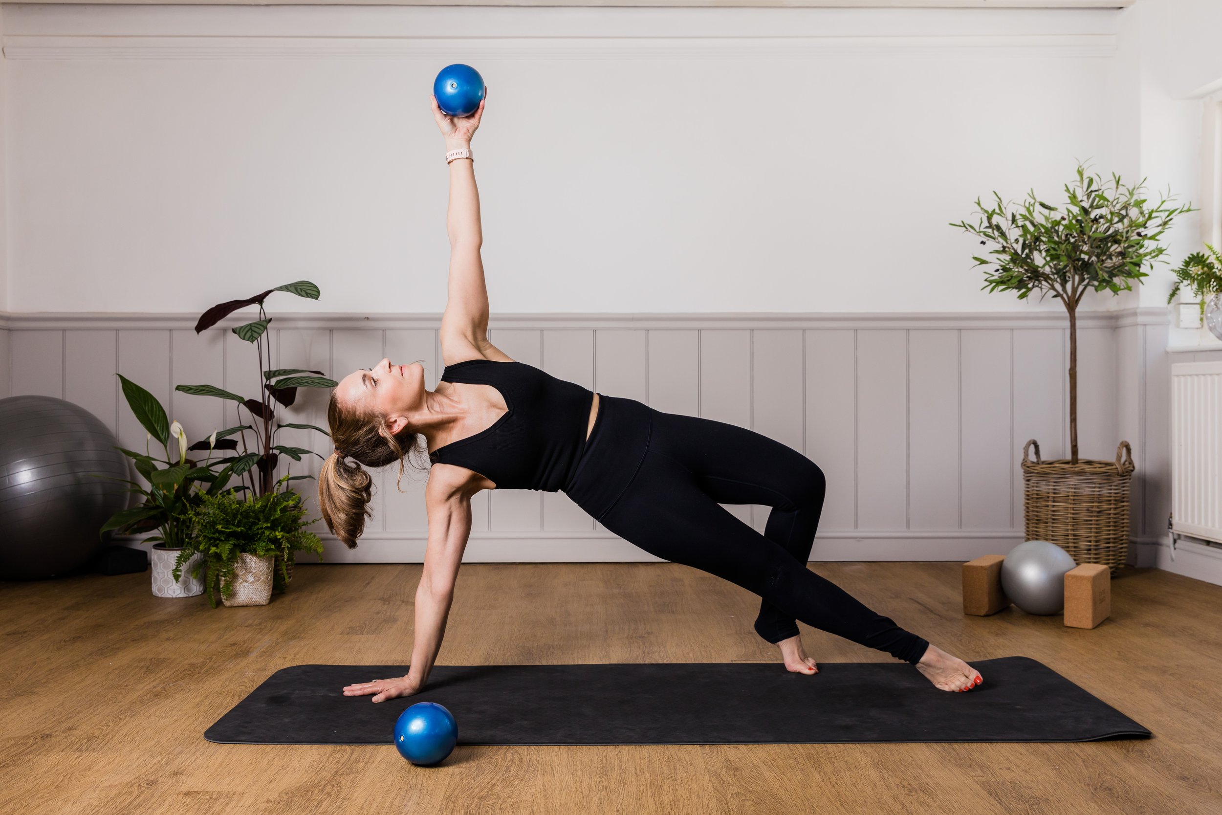 A woman performing a yoga pose in a room with plants and exercise equipment, holding a blue exercise ball upright with one hand while balancing on the opposite arm and foot.
