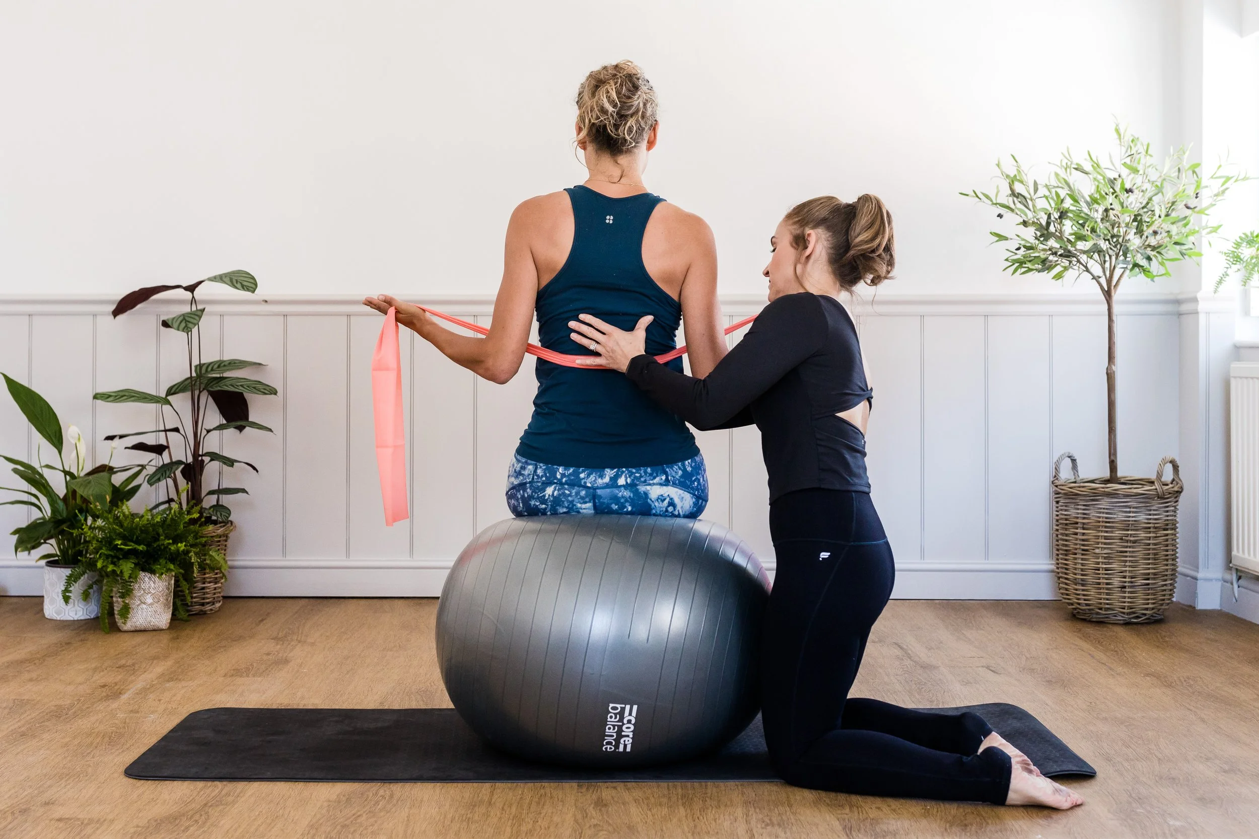 A woman performs a physical therapy exercise with a resistance band while kneeling on an exercise ball, guided by a trainer in a bright room with plants nearby.