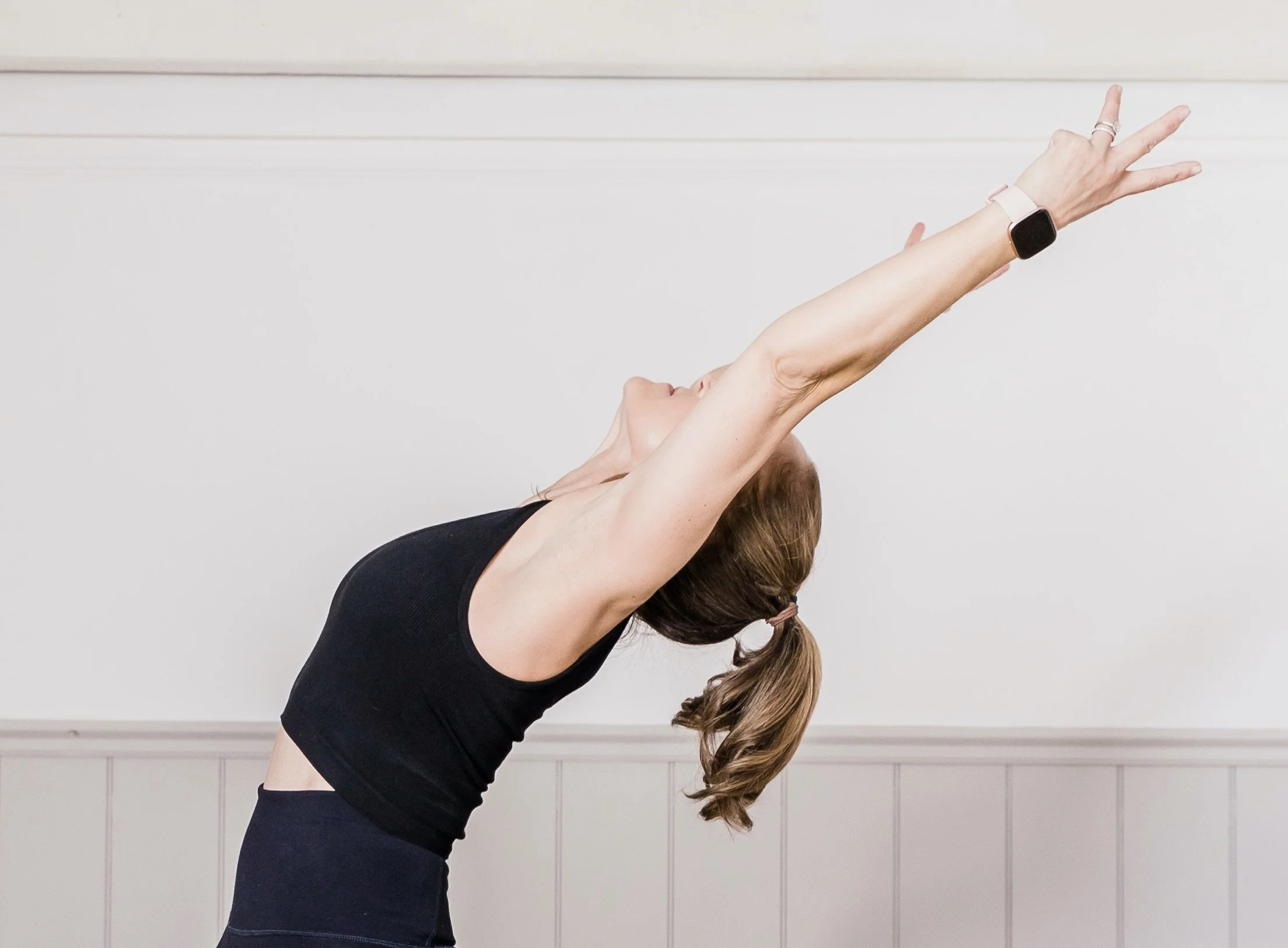 A woman with long braided hair is practicing yoga or stretching indoors. She's wearing a black sports bra and black leggings, with a smartwatch on her left wrist. She is extending her arms upwards and outwards, leaning back slightly.