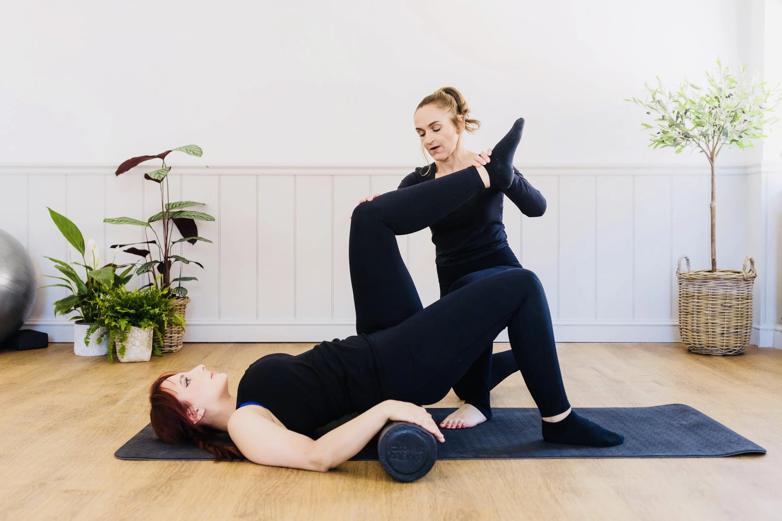 Two women practicing acro yoga indoors, one lying on her back on a yoga mat with a black foam roller in her hand, the other kneeling on her partner's legs with her hands on her partner's knee and shoulder, surrounded by houseplants and a white wall.