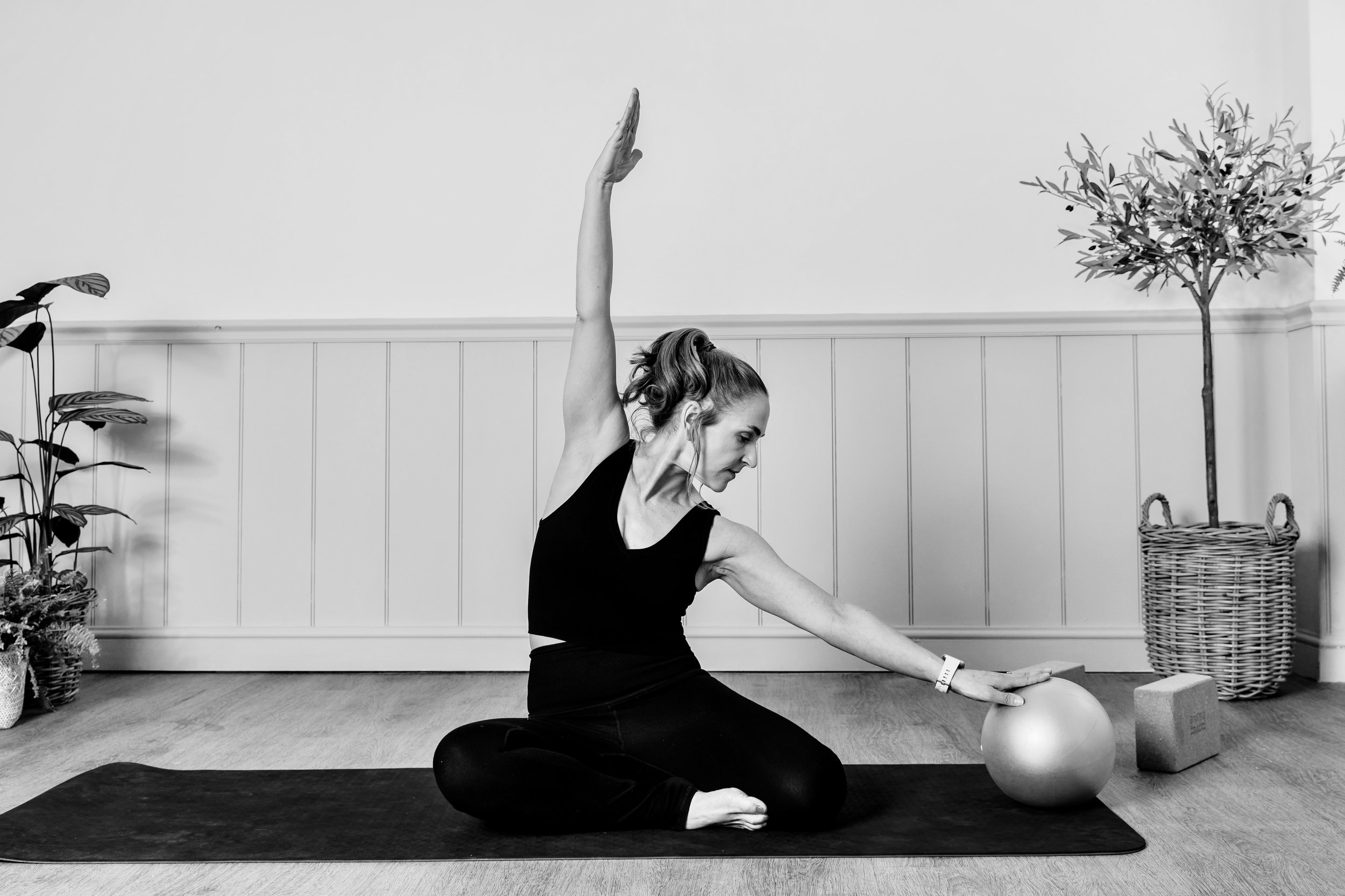 A woman practicing yoga indoors in a seated stretch pose, extending one arm upward and the other arm to the side, on a yoga mat, with plants and a basket in the background.