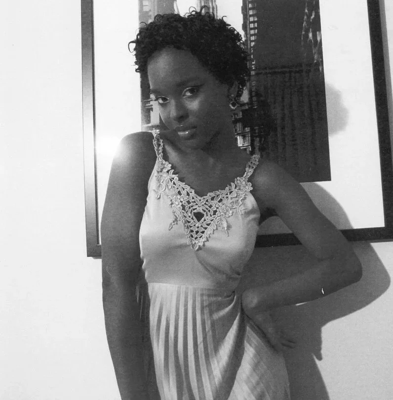 A Black woman with short curly hair wearing a sleeveless top with lace detail and a striped skirt, standing indoors in front of framed artwork.