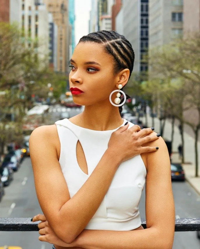 A woman with braided hair, wearing a white sleeveless top and large earrings, standing outdoors on a balcony or rooftop in an urban setting with tall buildings and trees in the background.