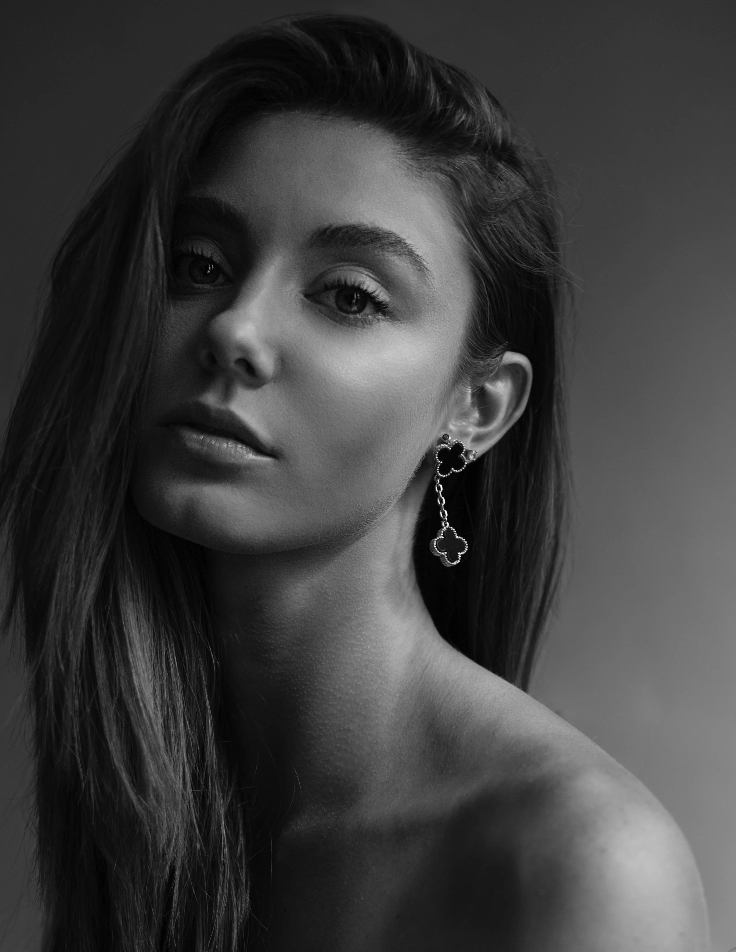 Close-up black and white portrait of a woman with long hair, jewelry, and makeup, with a neutral background.