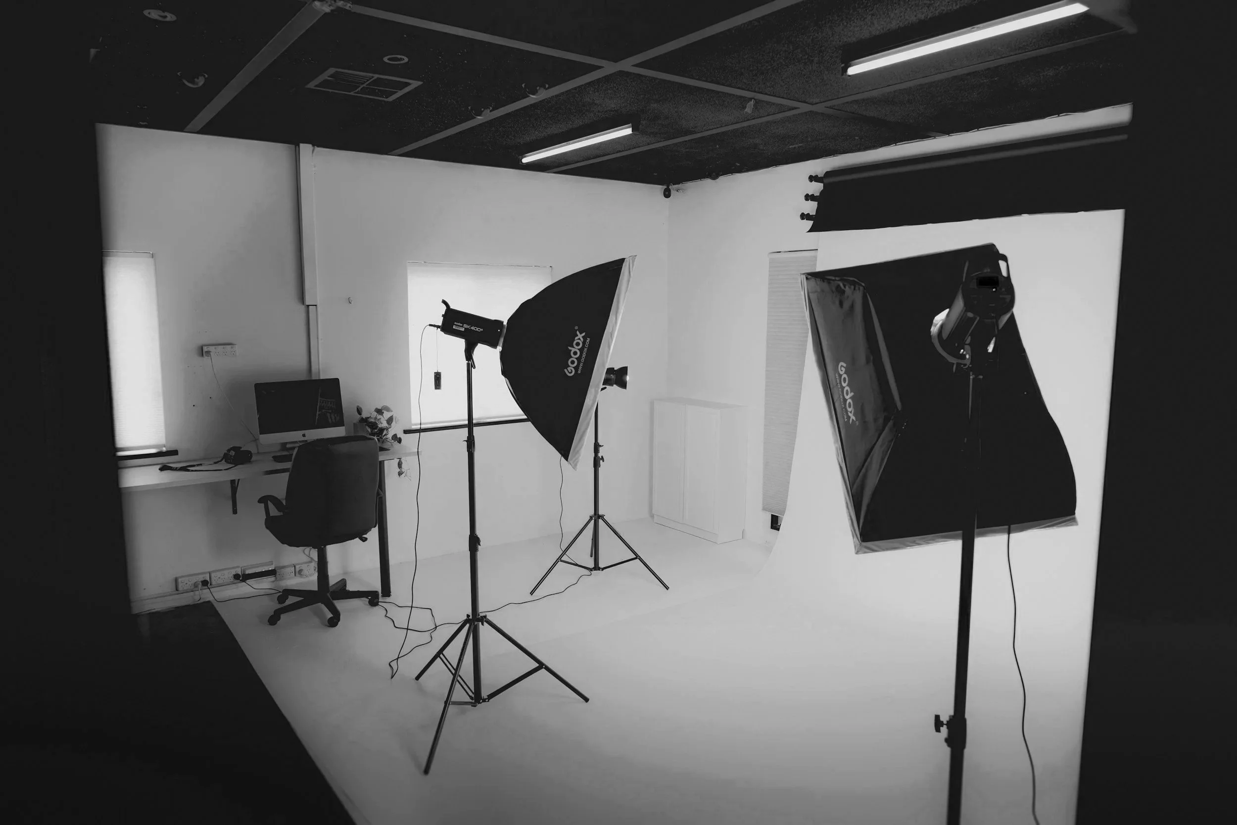 Photography studio setup with lighting equipment, backdrop, computer on desk, and an office chair.