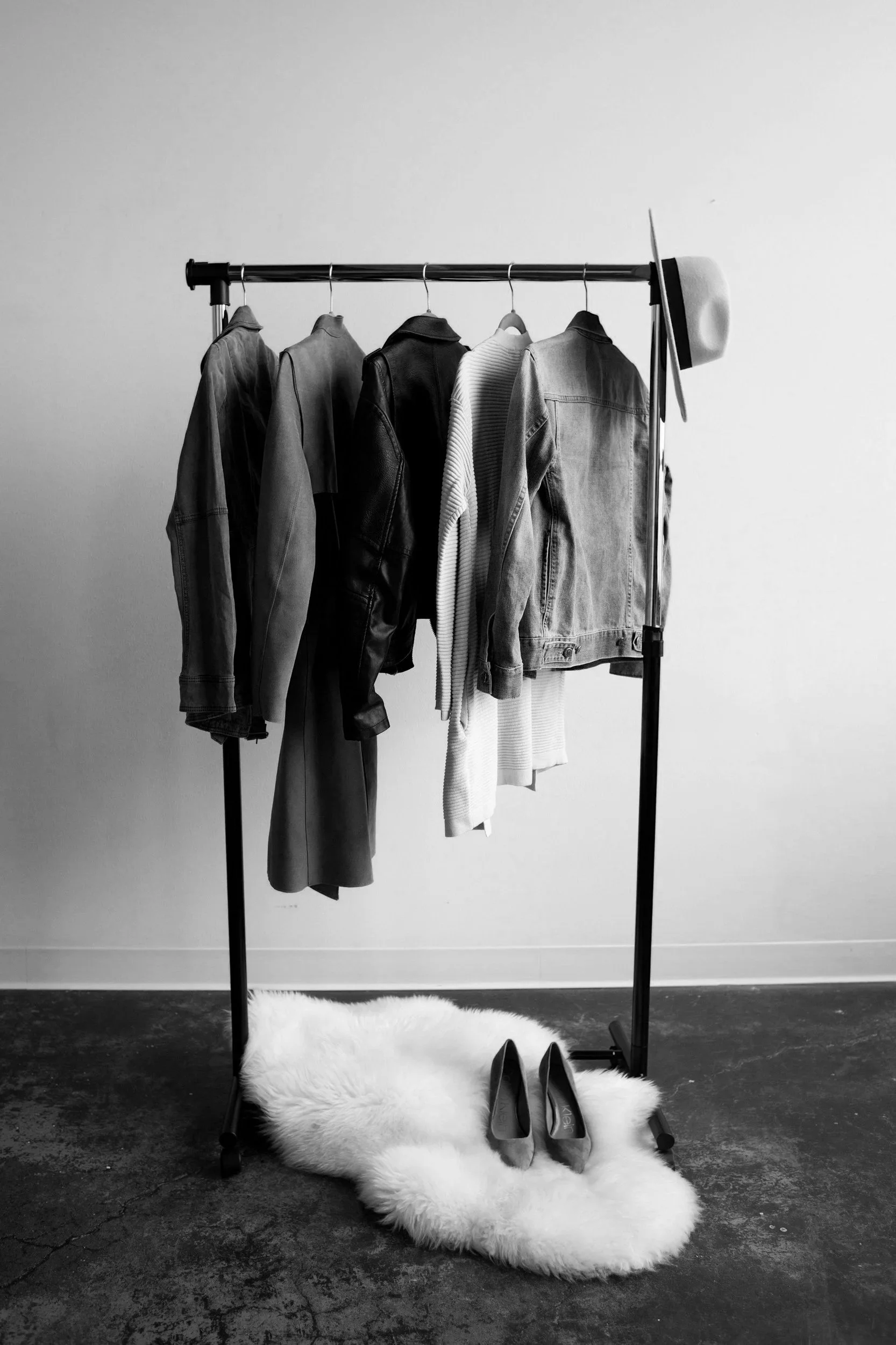 Black and white photograph of a clothing rack holding several jackets and shirts, with a white fedora hat hanging on the right side. The rack is positioned against a plain wall, and there is a fluffy white rug with a pair of high-heeled shoes on it on the floor.